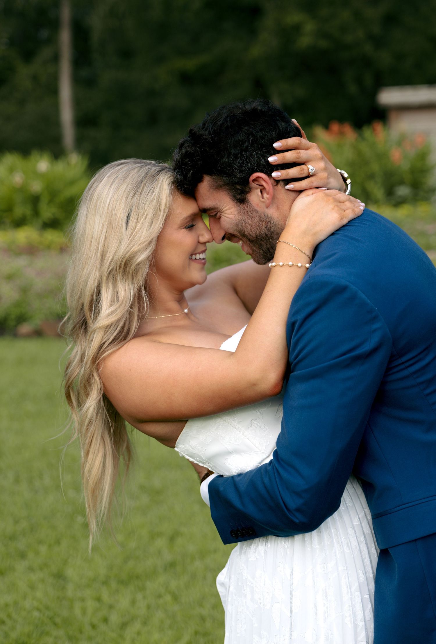 Romantic couple nearly kissing outdoors, bride in white dress and groom in navy suit embracing in garden.