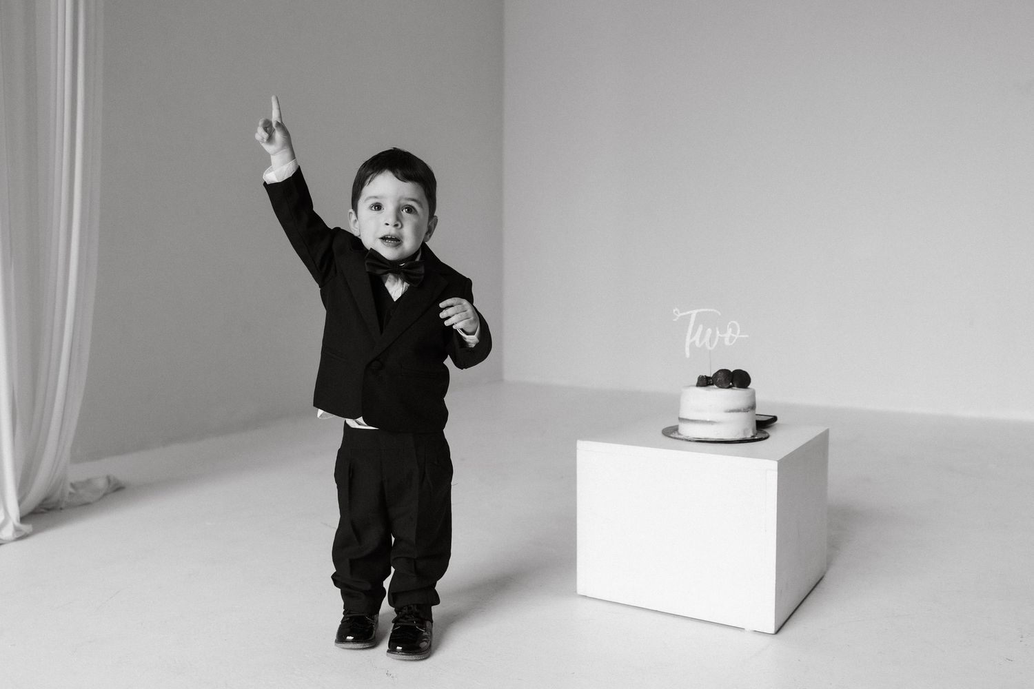 Toddler boy in black suit pointing up beside birthday cake with two topper, black and white photo.