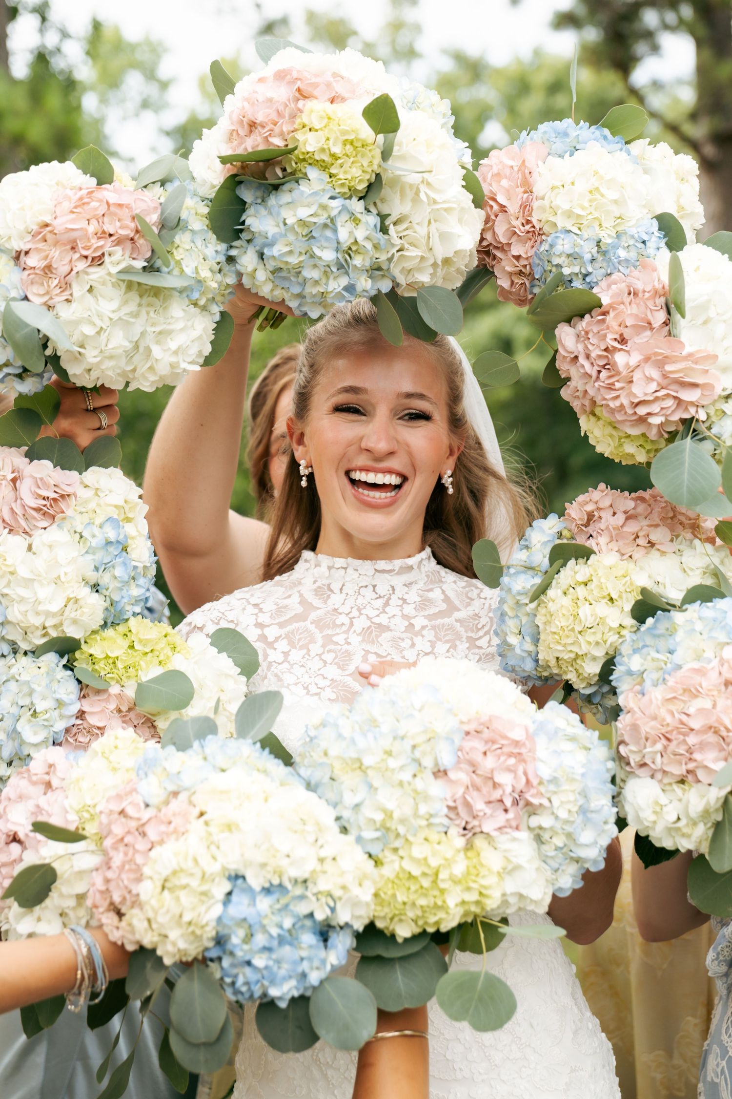 Smiling bride surrounded by bridesmaids holding pastel pink, blue, and white hydrangea bouquets at outdoor wedding.