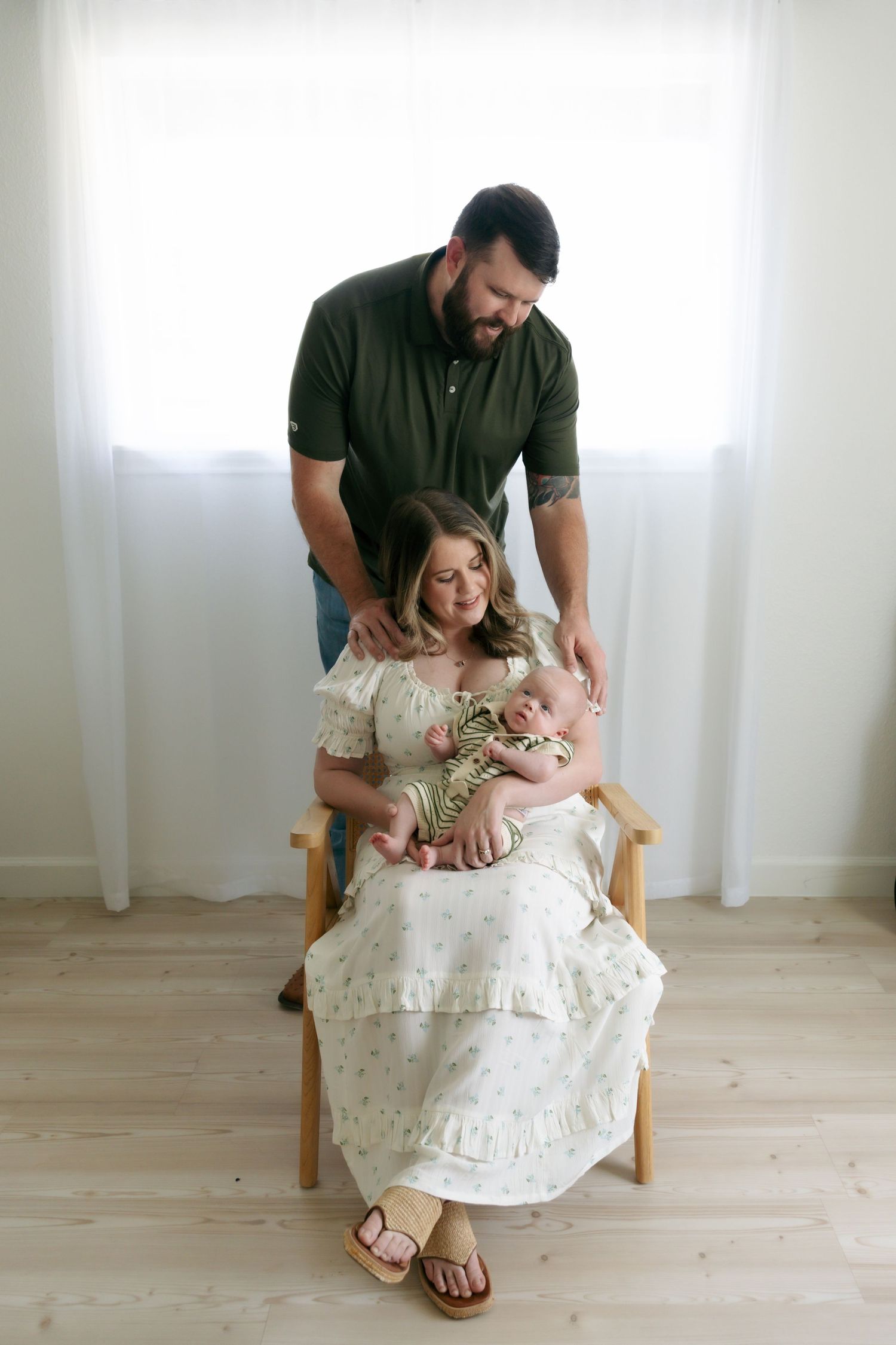 New parents with newborn baby, mother seated in chair holding infant while father stands behind.