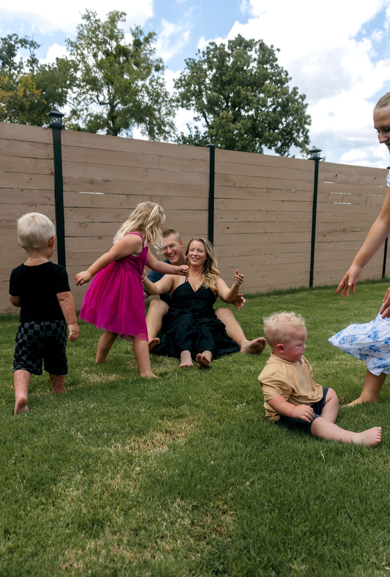 Family sitting and playing together on green grass in a backyard with a wooden fence.