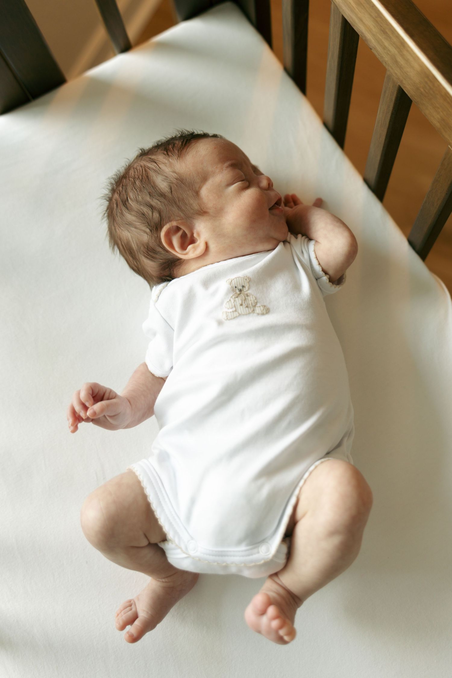 Newborn baby sleeping peacefully on back in a white crib wearing a white onesie.