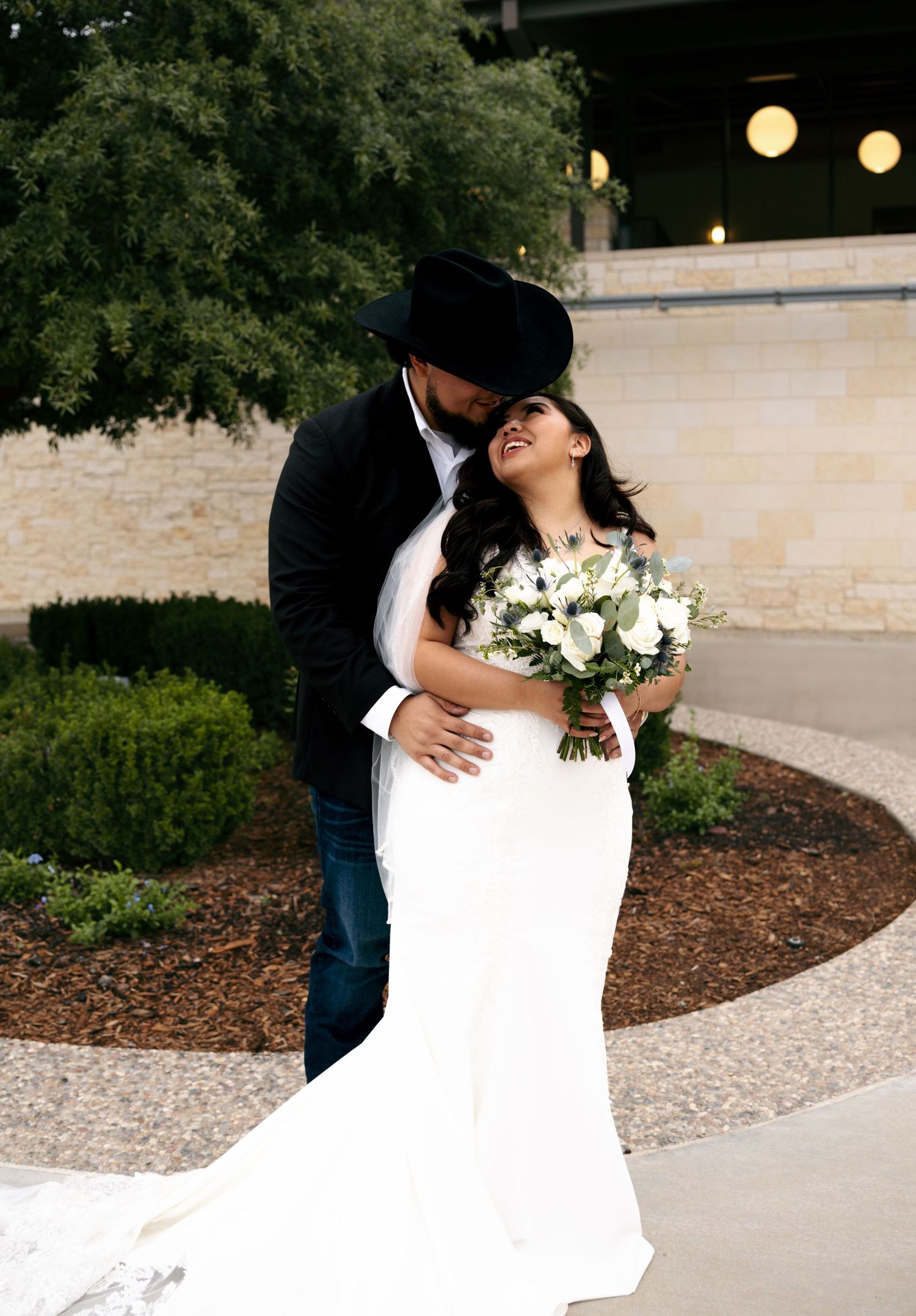 Bride and groom sharing romantic kiss outdoors, groom wearing black cowboy hat.