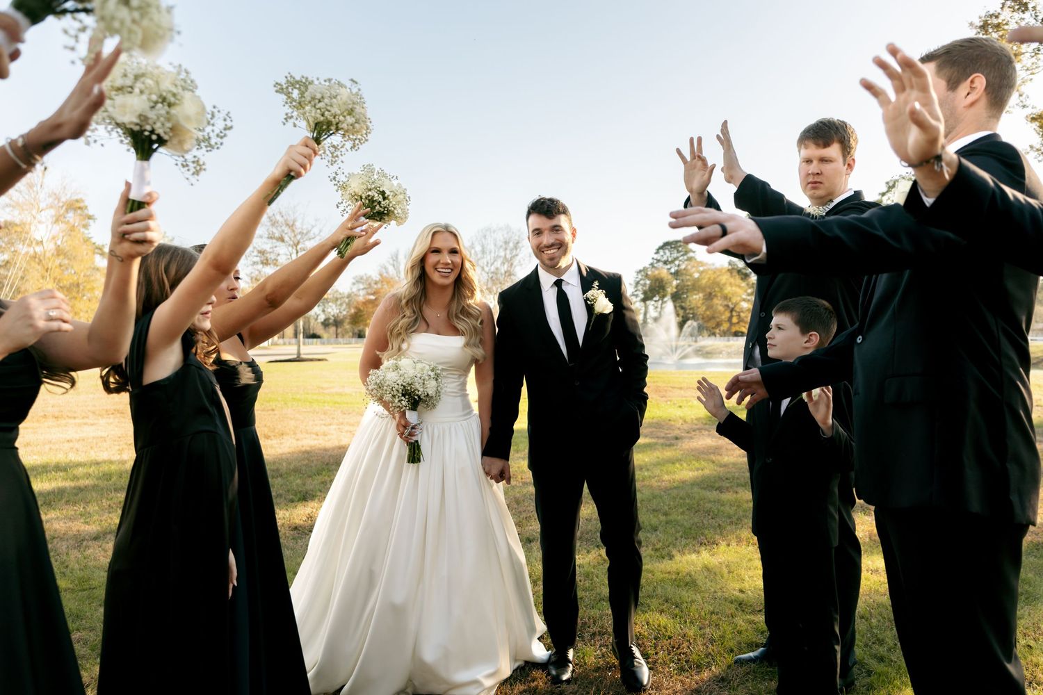 Wedding party in black attire celebrates with bride in white ball gown and groom on sunny outdoor lawn.