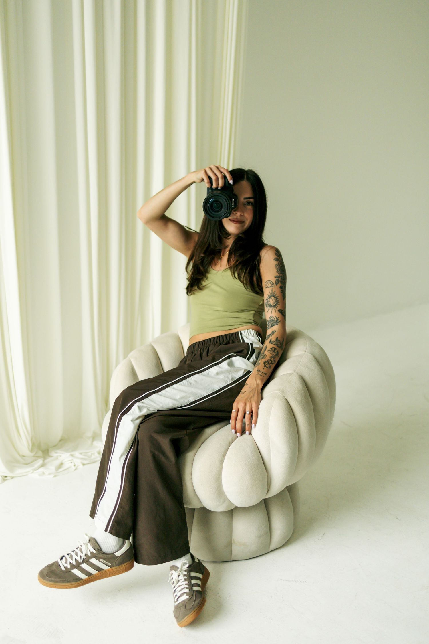 Tattooed female photographer in green top sitting in white chair holding Canon camera in bright studio.