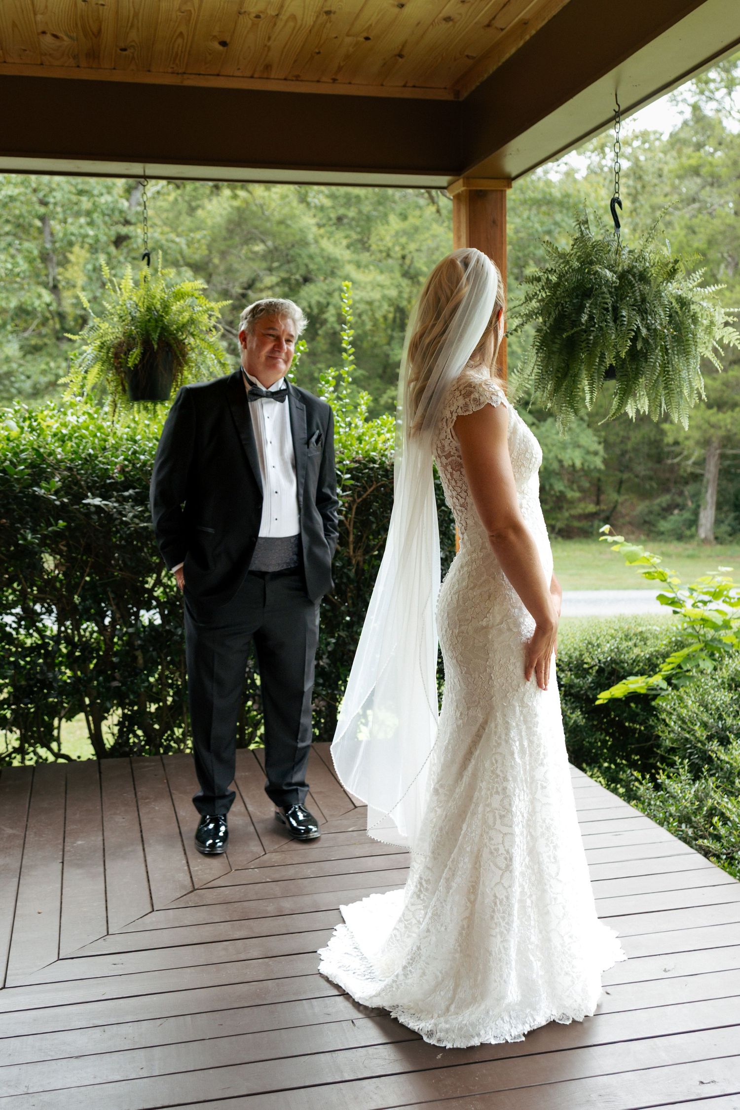 Bride in lace wedding gown with veil stands on porch as groom in tuxedo admires her amid lush greenery.