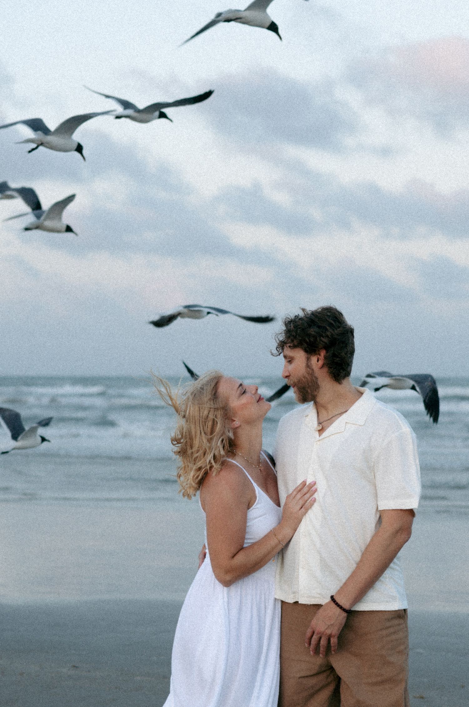 Romantic couple in white embracing on beach with seagulls flying overhead at dusk.