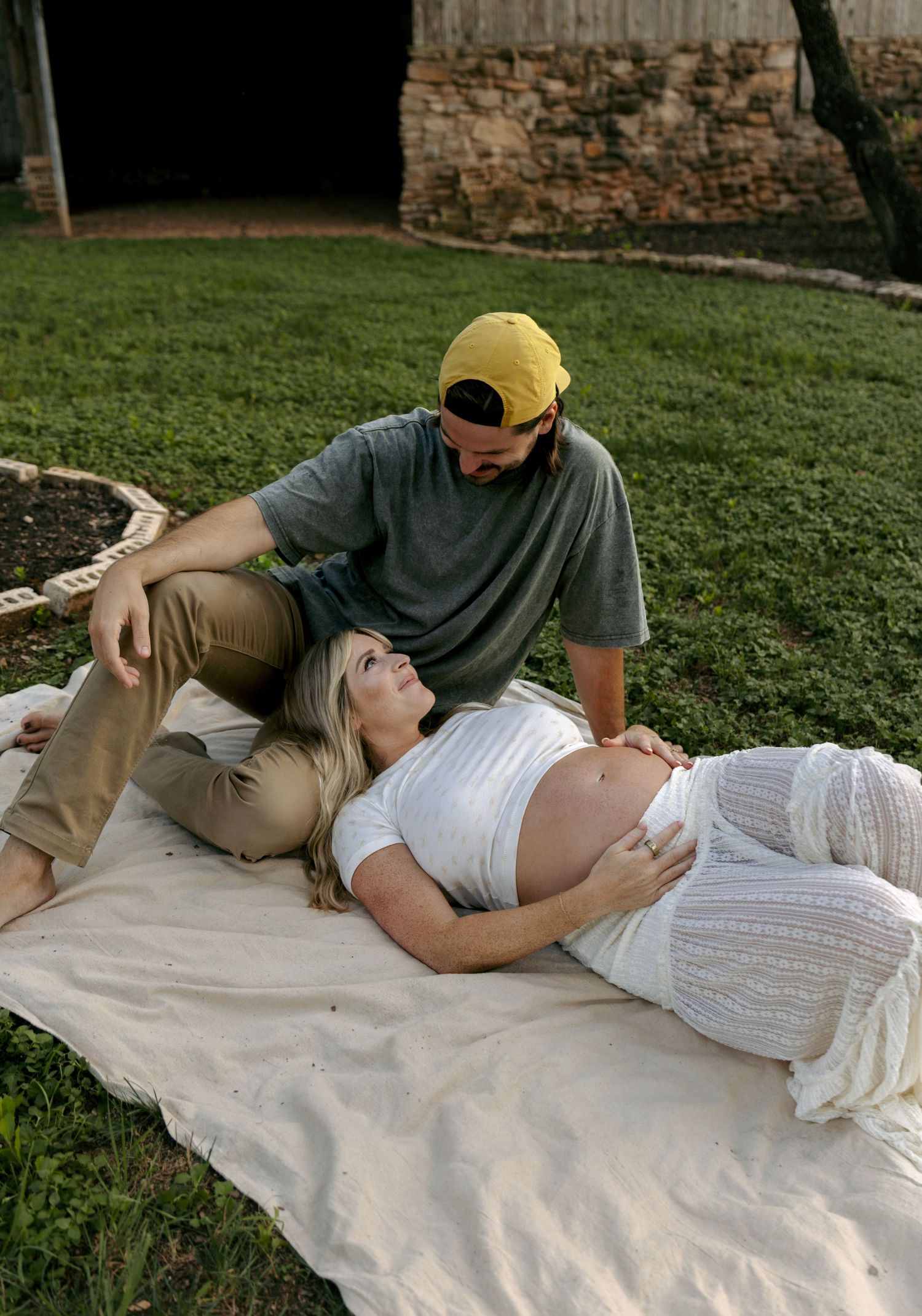 Pregnant woman in white dress lying on blanket outdoors while partner in yellow cap sits beside her.