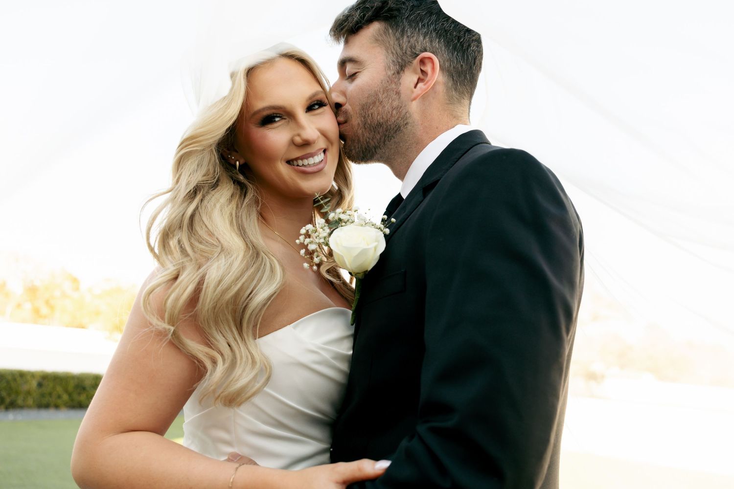 Groom in black tuxedo kisses bride in white strapless gown on cheek under white tent at outdoor wedding.
