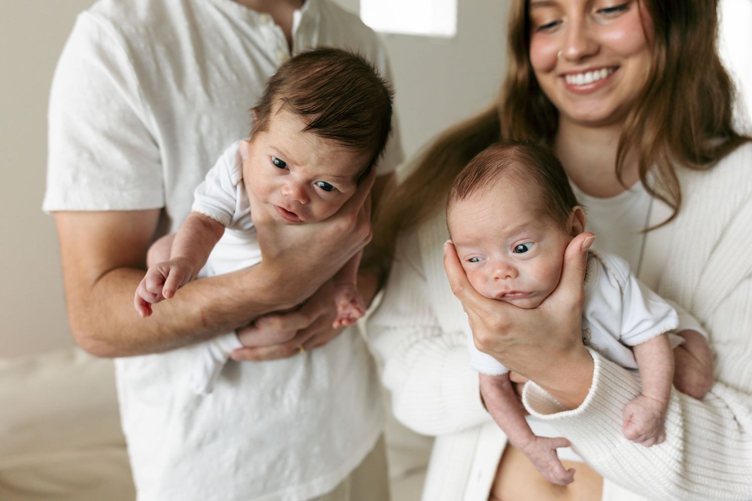 Parents hold two newborn twins side by side, both babies alert and looking forward in bright room.