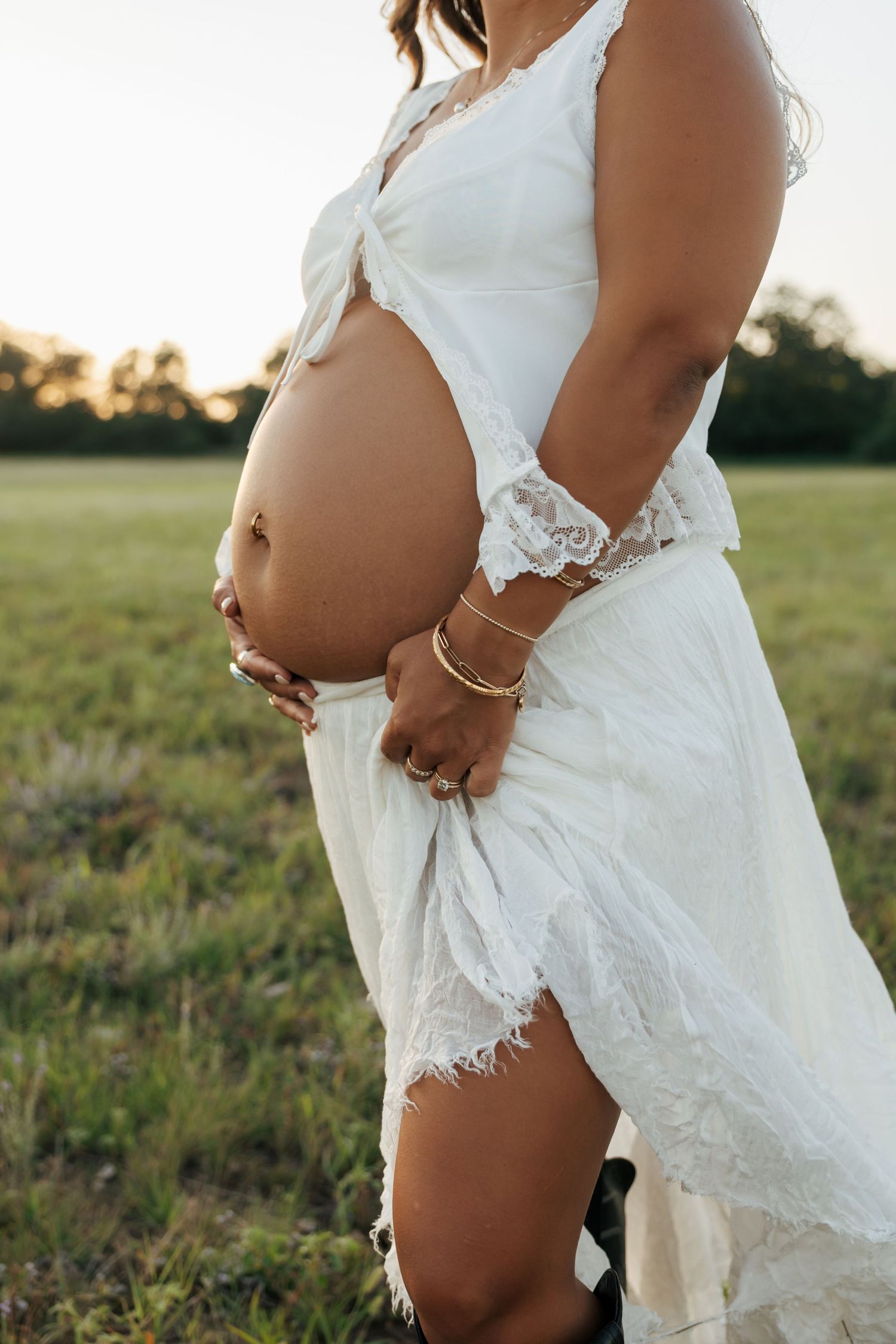 Close-up of a pregnant woman in a white lace dress cradling her bare baby bump outdoors.