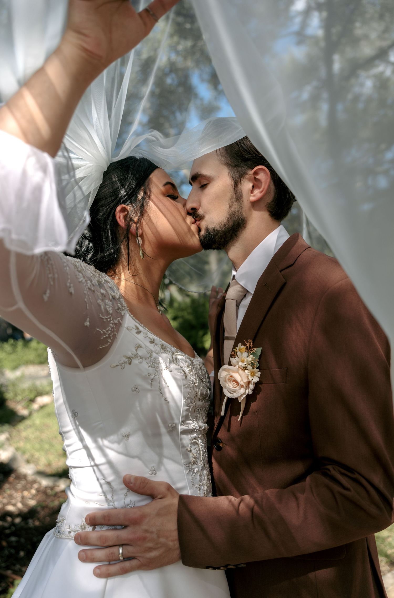 Bride and groom sharing romantic kiss under flowing white veil outdoors on their wedding day.