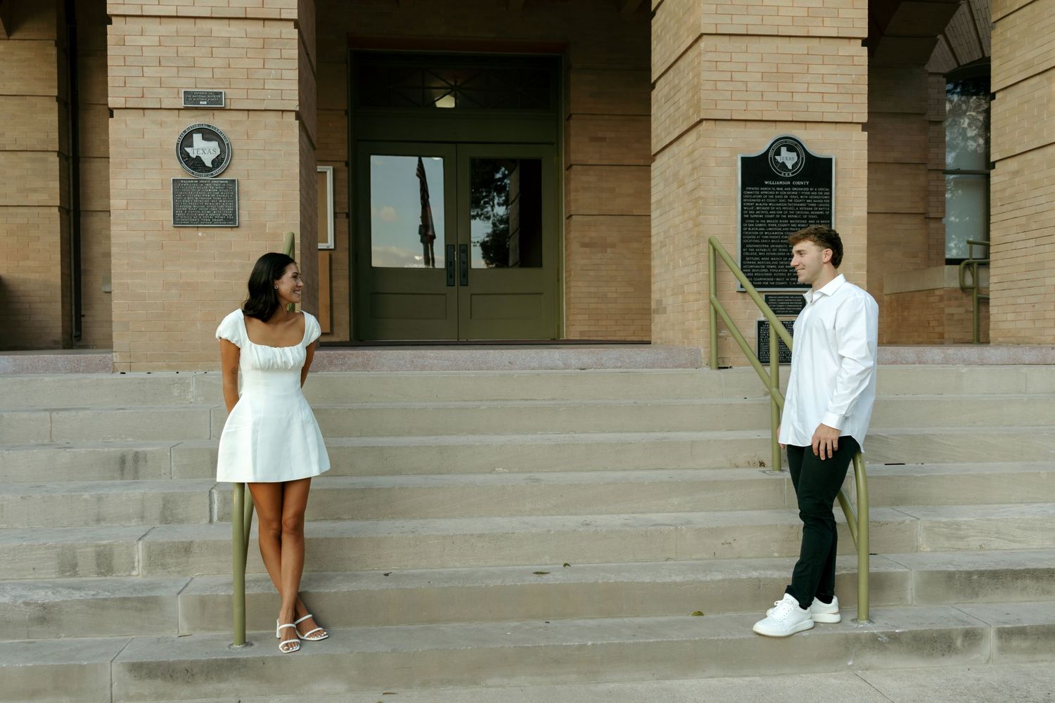 Couple standing apart on steps of a historic brick building, smiling at each other.
