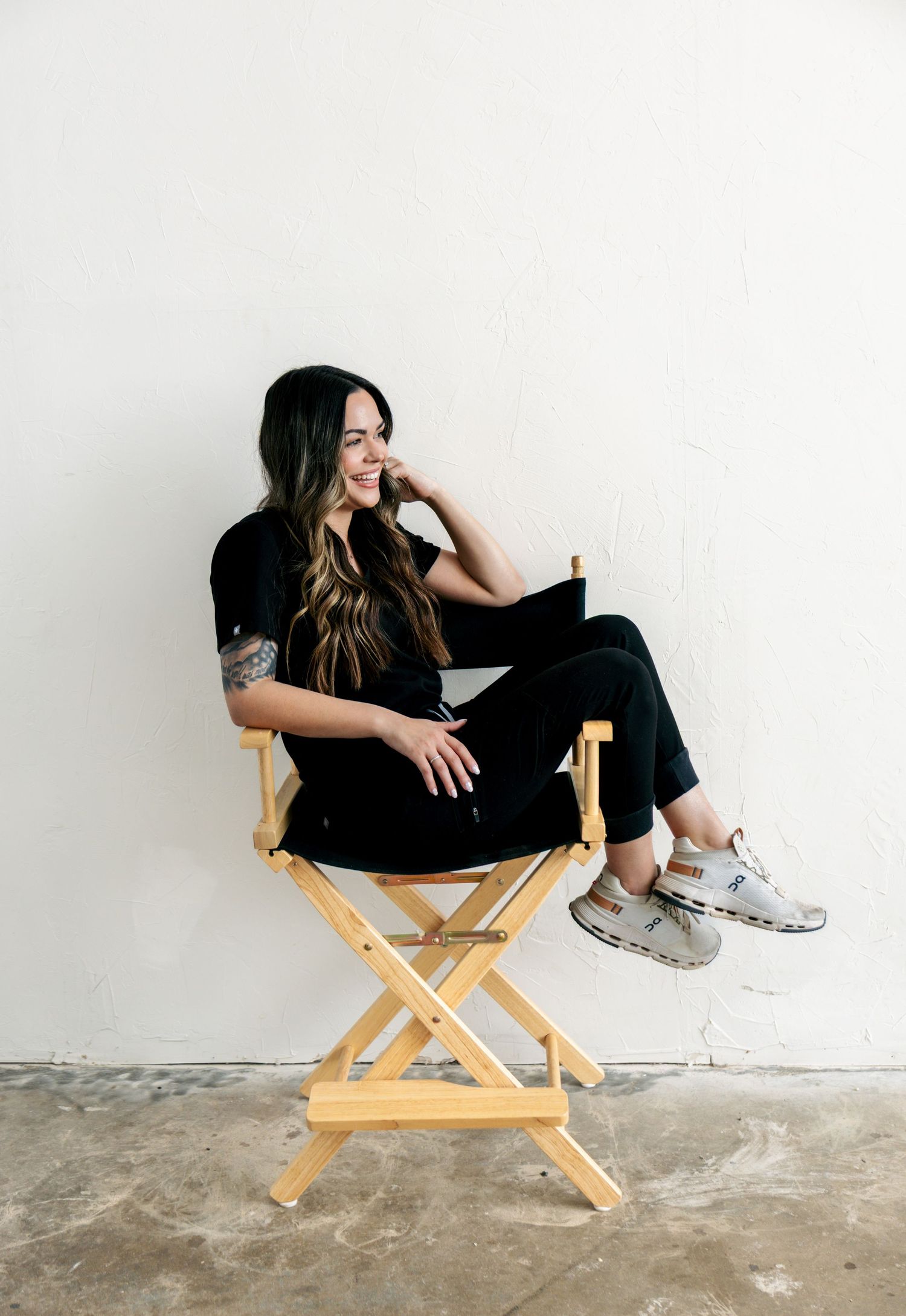 Woman in black outfit laughing while seated in wooden directors chair against white wall.