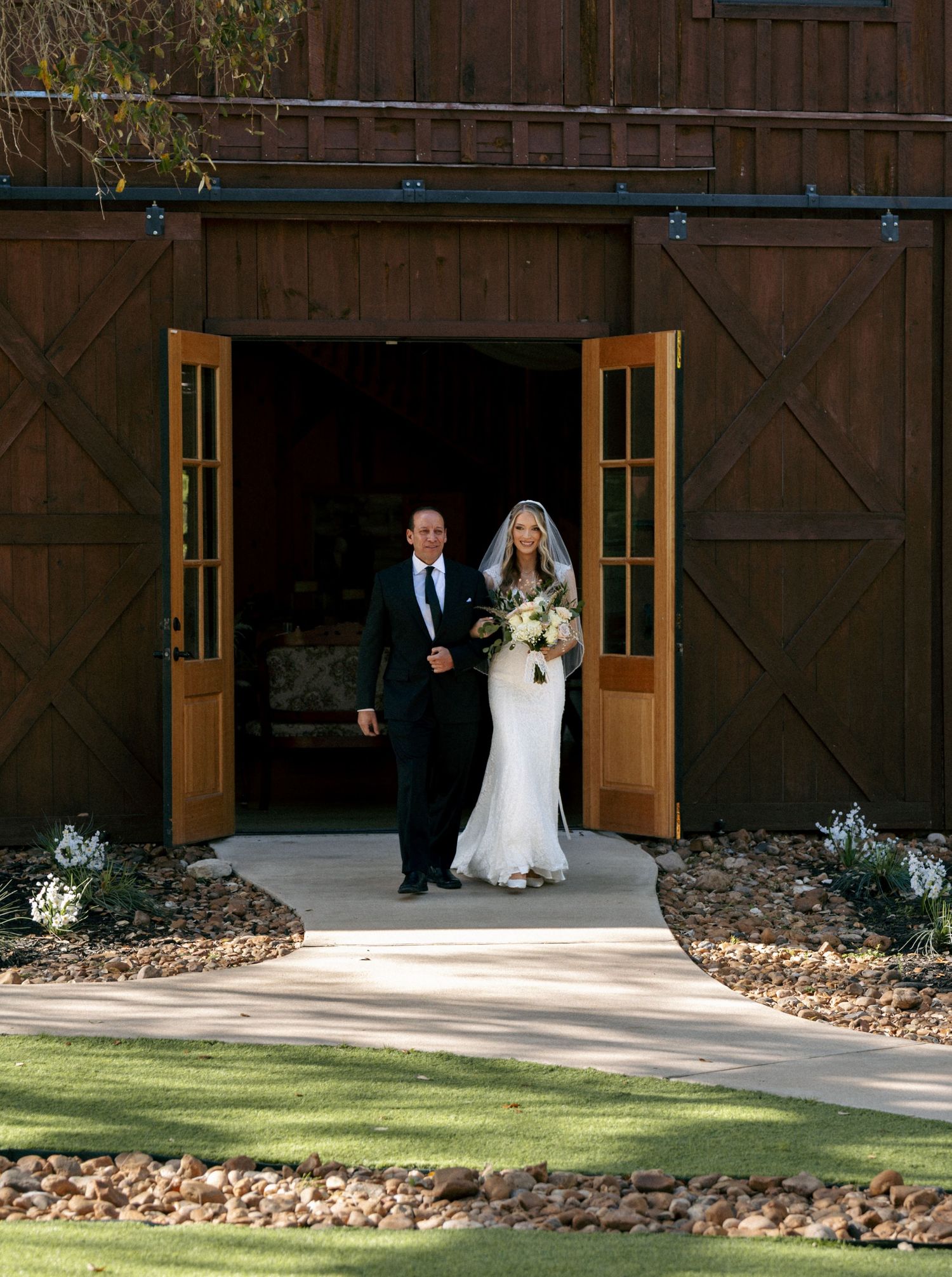 Bride in white gown with veil and escort exit rustic wooden barn doors on wedding day.