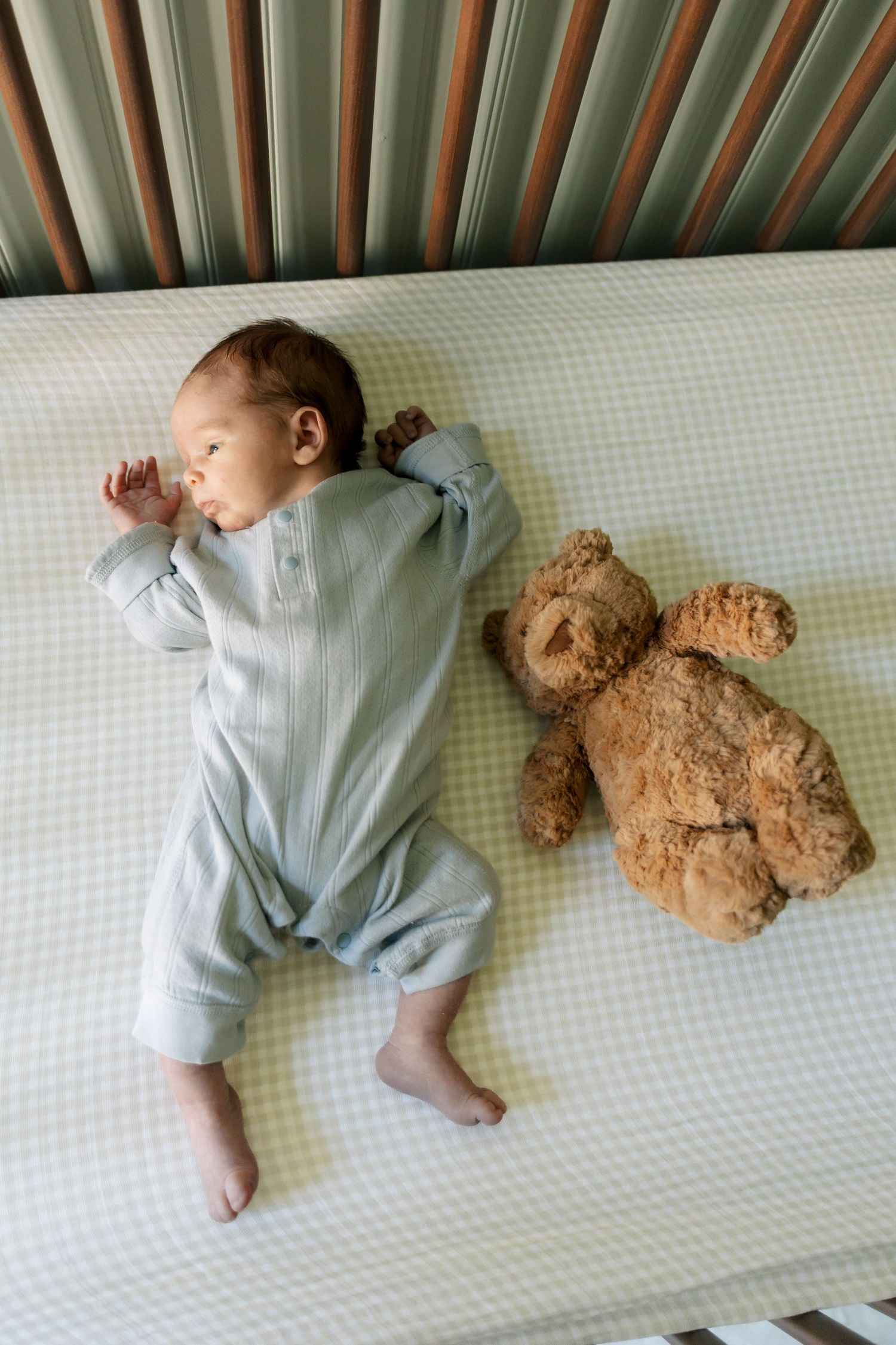 Newborn baby sleeping peacefully in a crib wearing a light blue romper beside a brown teddy bear.