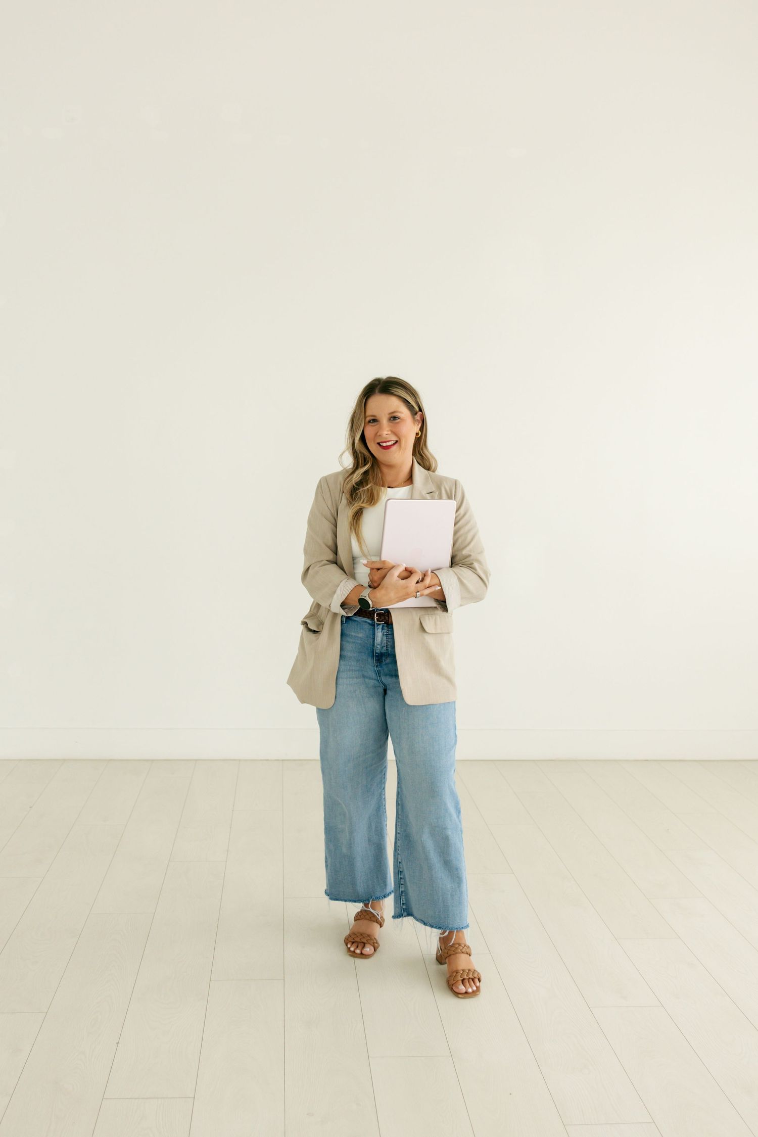 Smiling blonde woman in beige blazer holding notebook and coffee cup against clean white studio wall.