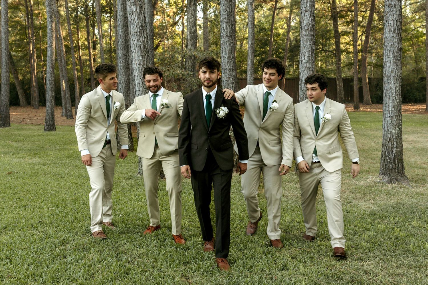 Groom in black suit walking confidently with four groomsmen in tan suits through a pine forest.