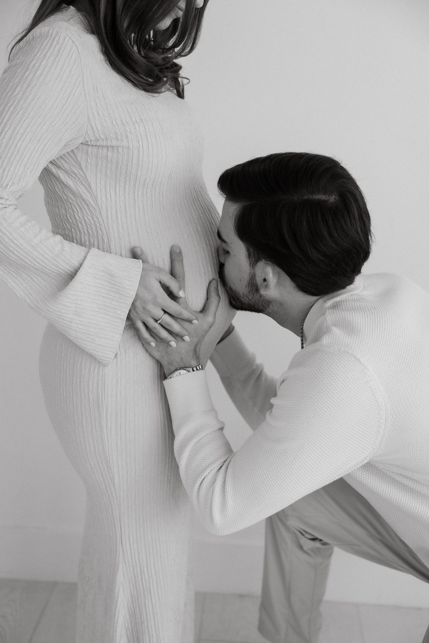 Man kneeling and kissing pregnant womans baby bump in white dress, black and white maternity portrait.