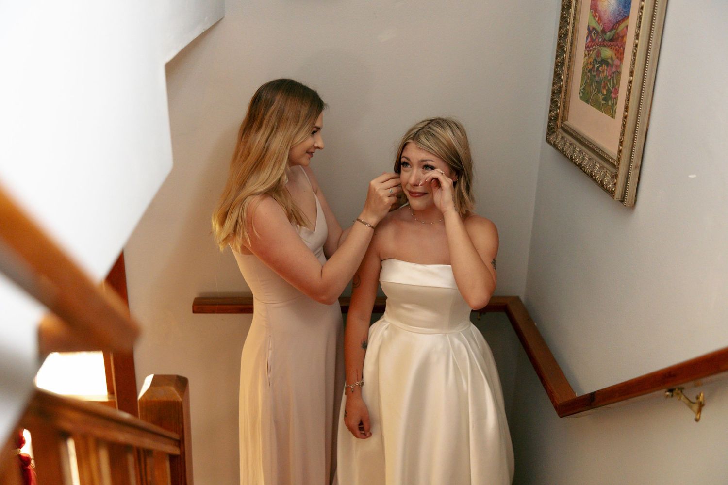 Bridesmaid helping emotional bride wipe tears on staircase before wedding ceremony indoors.