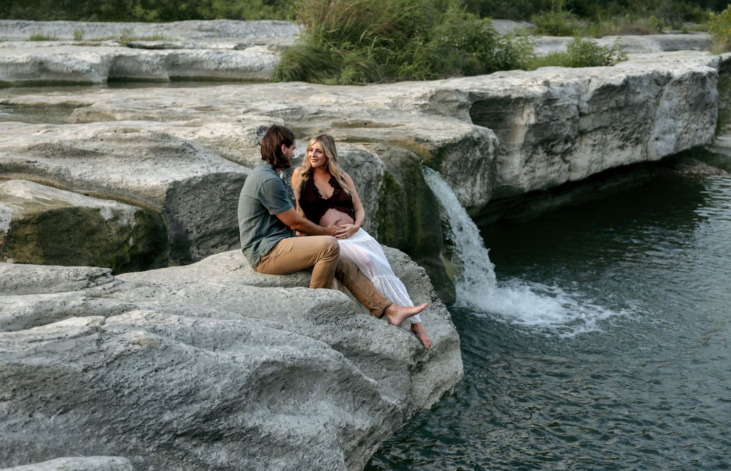Couple sitting together on rocks beside a natural waterfall in an outdoor setting.
