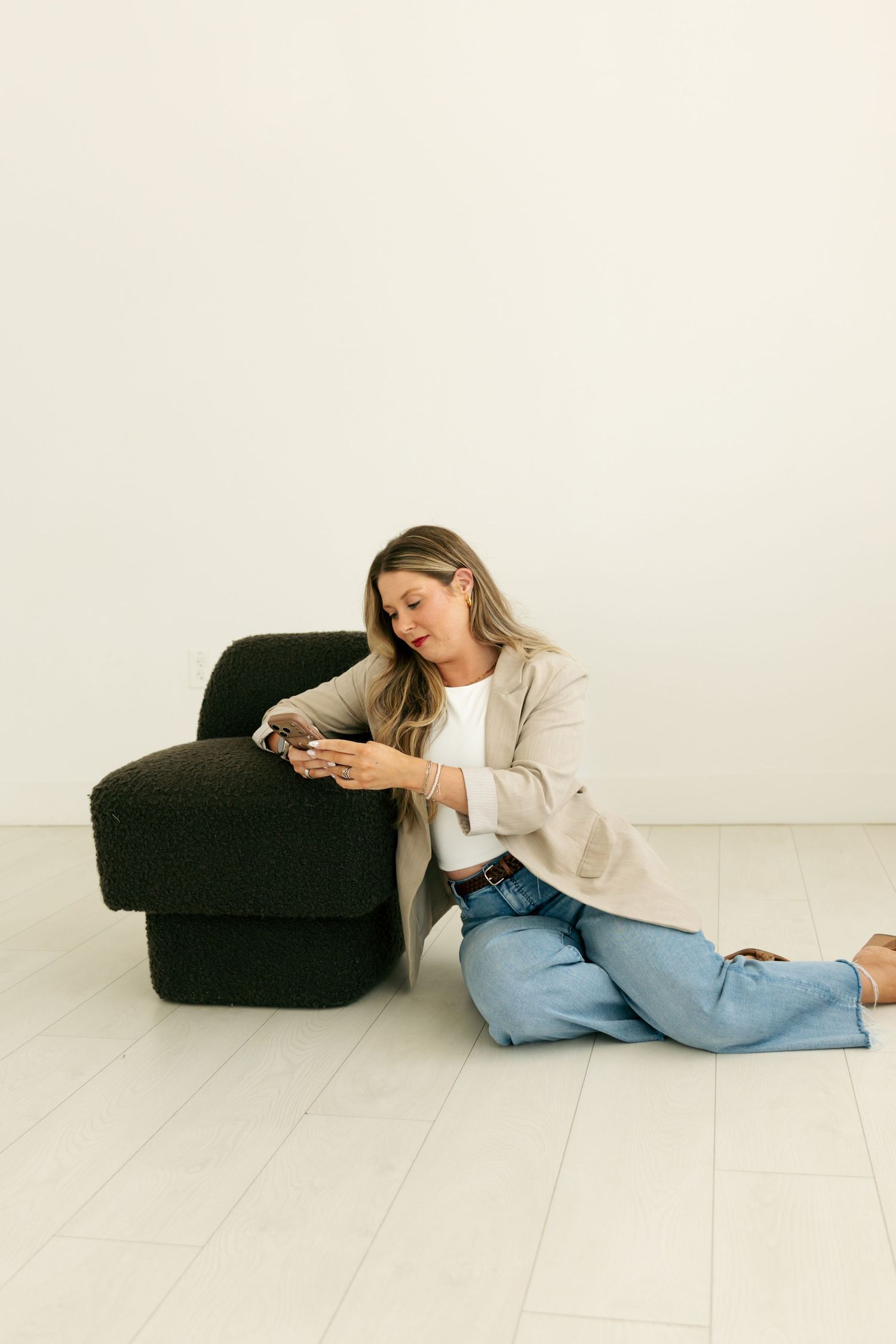 Blonde woman in blazer and jeans sitting on floor using smartphone next to black boucle chair.
