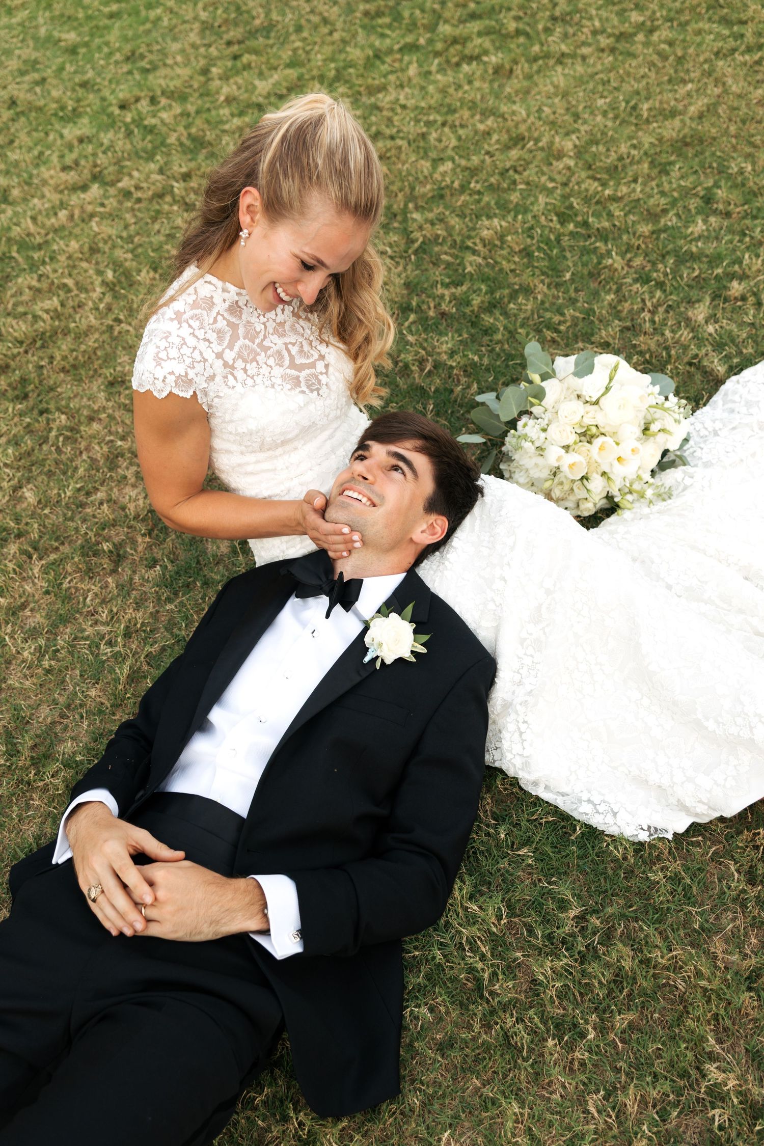 A smiling bride in white lace gown sits beside her tuxedo-clad groom lying on green grass, holding hands at their wedding.