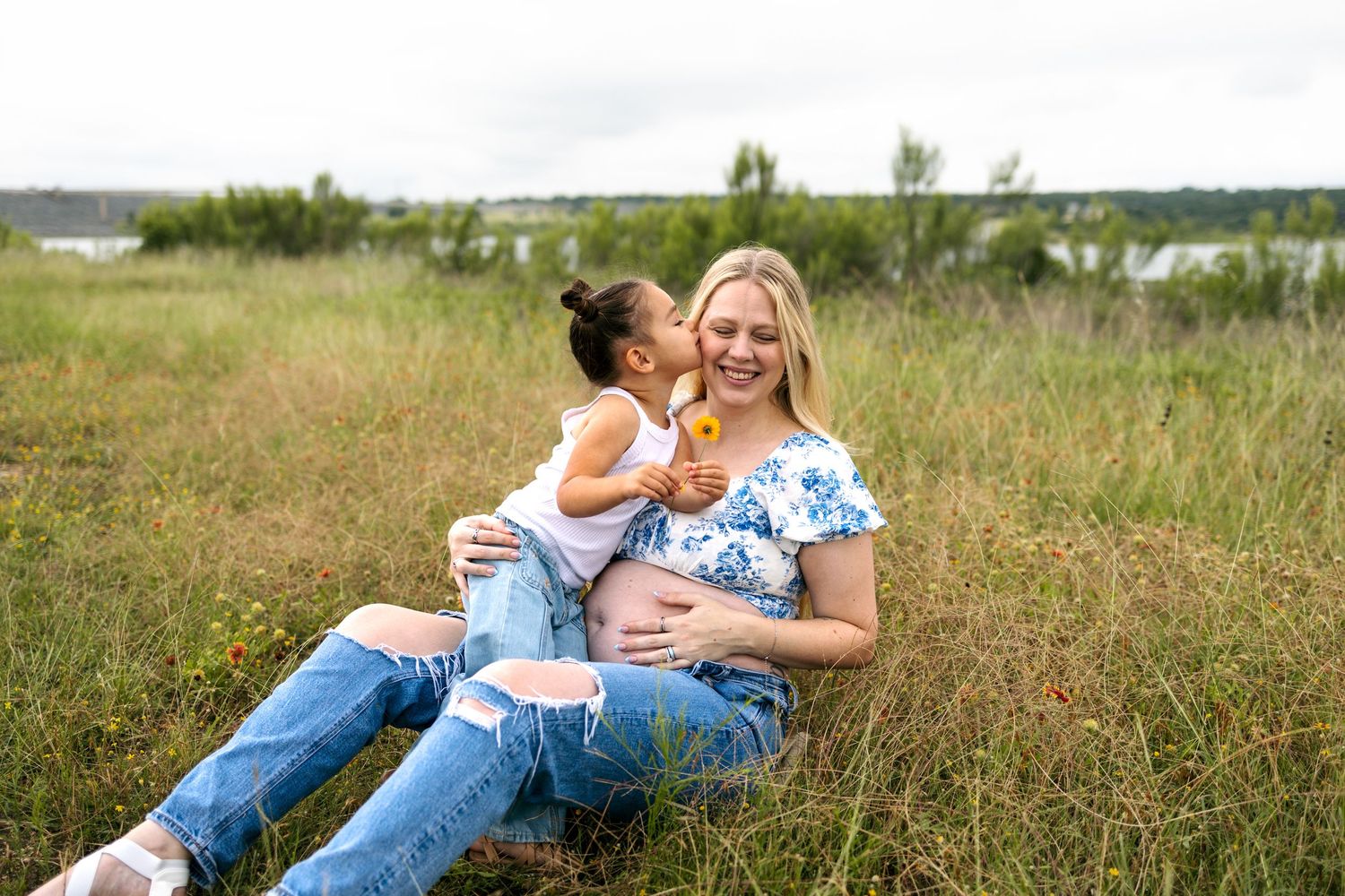 Pregnant woman sitting in green field with young girl kissing her cheek affectionately.