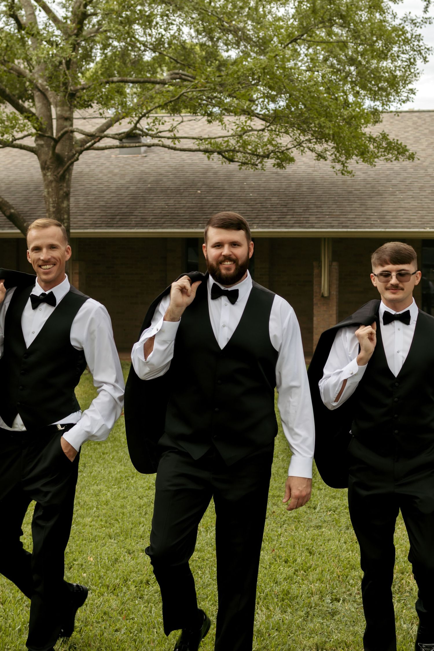 Groom and groomsmen walk confidently in black tuxedo vests and bow ties across a green lawn.