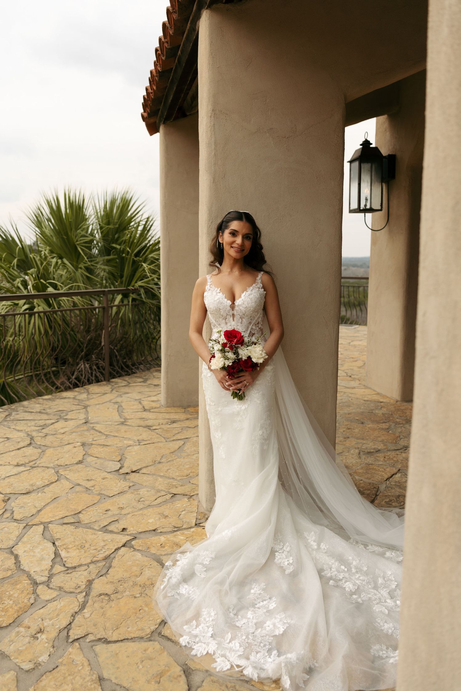 Elegant bride in lace mermaid gown holds red and white bouquet while posing at an outdoor wedding venue.
