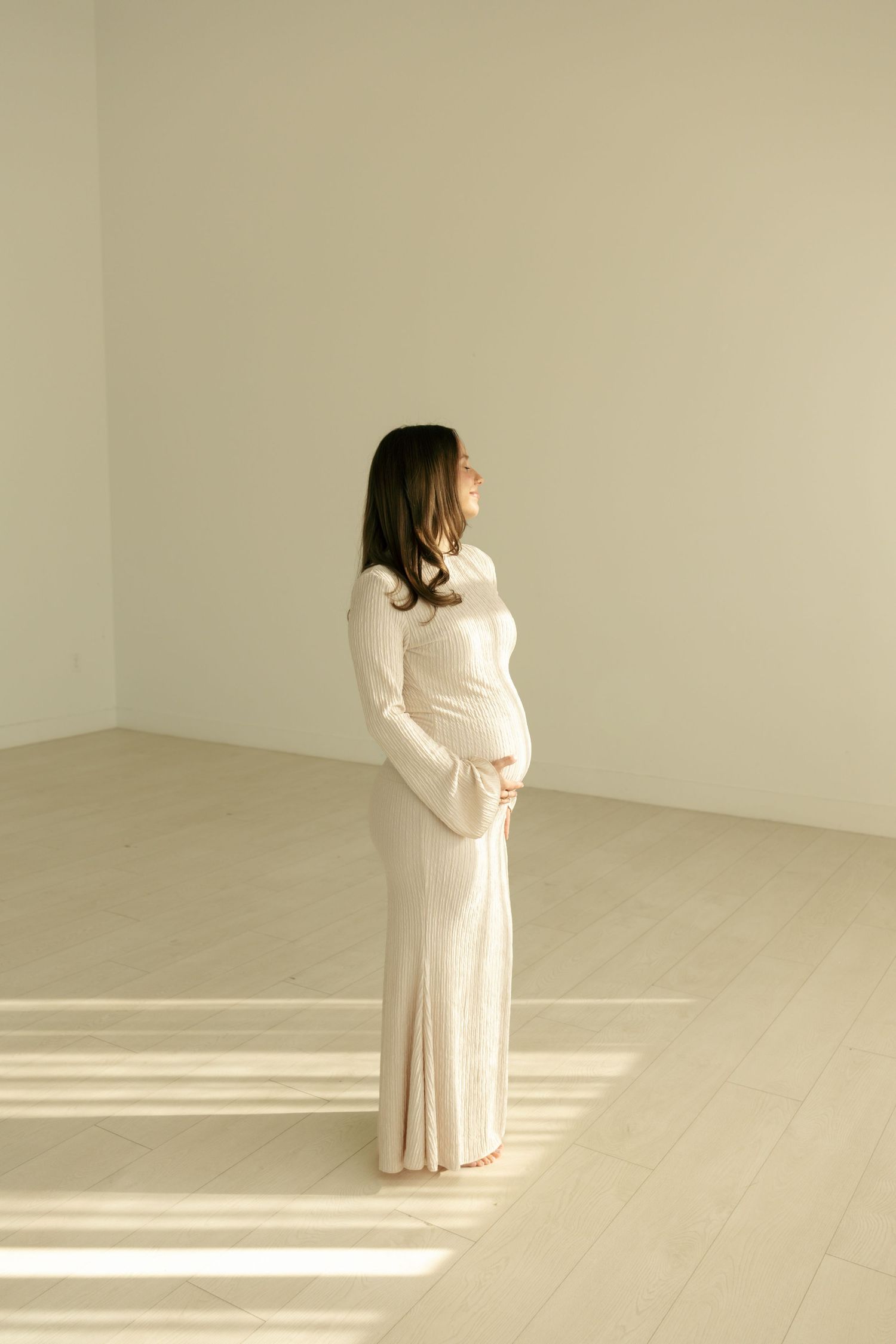 Pregnant woman in elegant cream maxi dress cradling baby bump in minimalist white studio with natural light.