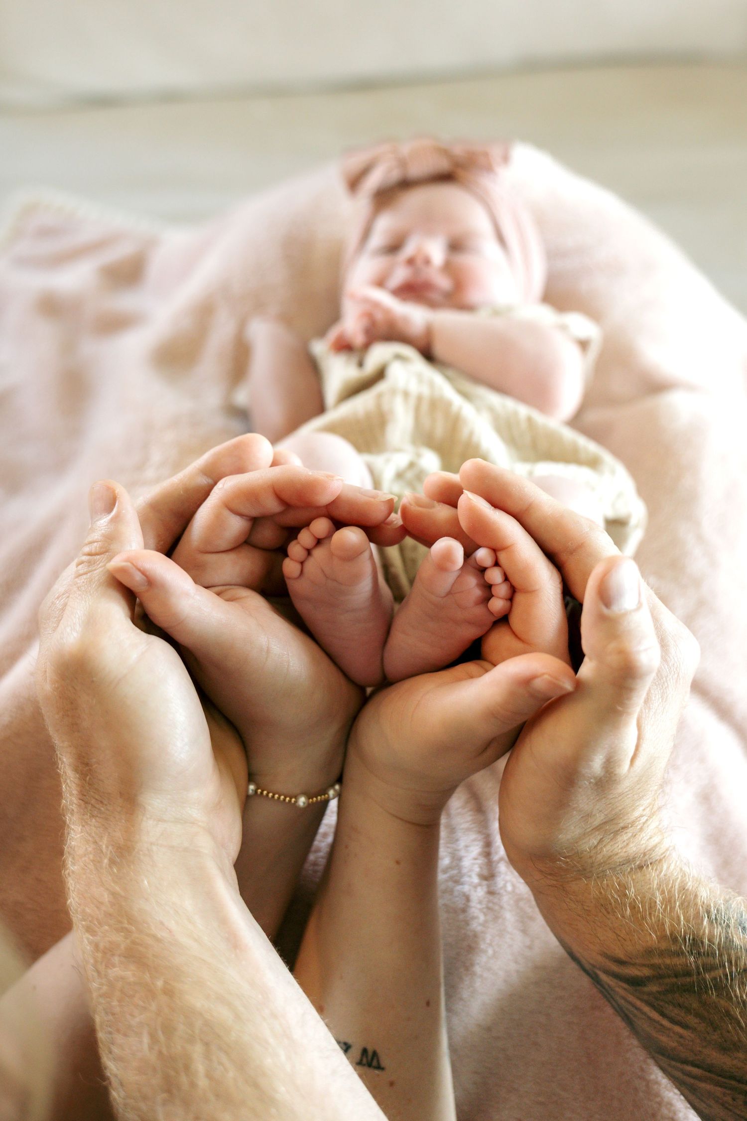 Parents cupping newborn baby's tiny feet in their hands on a pink blanket.
