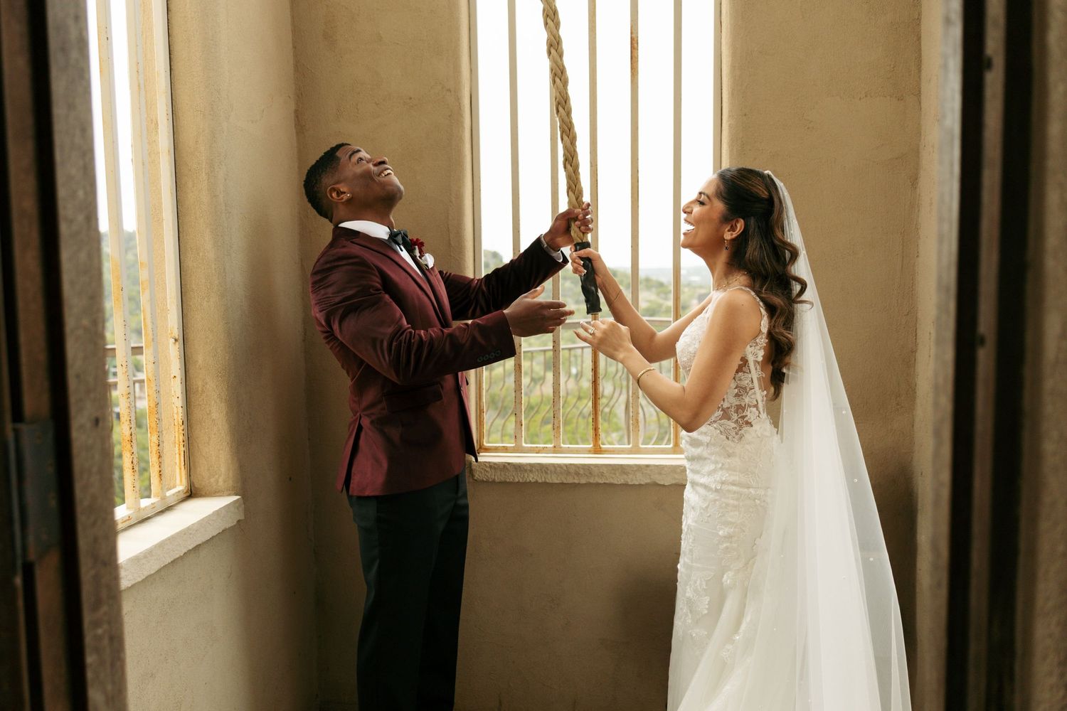 Newlywed couple playfully pulls a rope together inside a rustic wedding venue with warm natural lighting.