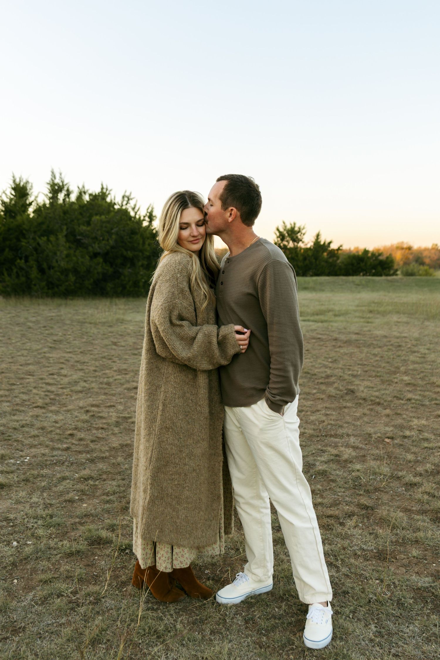 Man kissing woman on cheek at sunset, both standing in a field with trees.