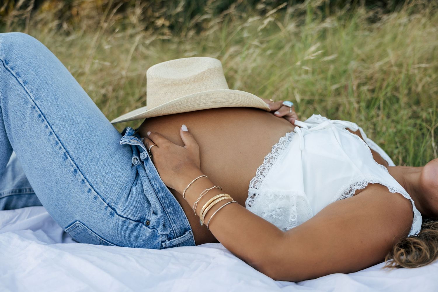 Pregnant woman lying in field wearing jeans and white top with cowboy hat resting on baby bump.