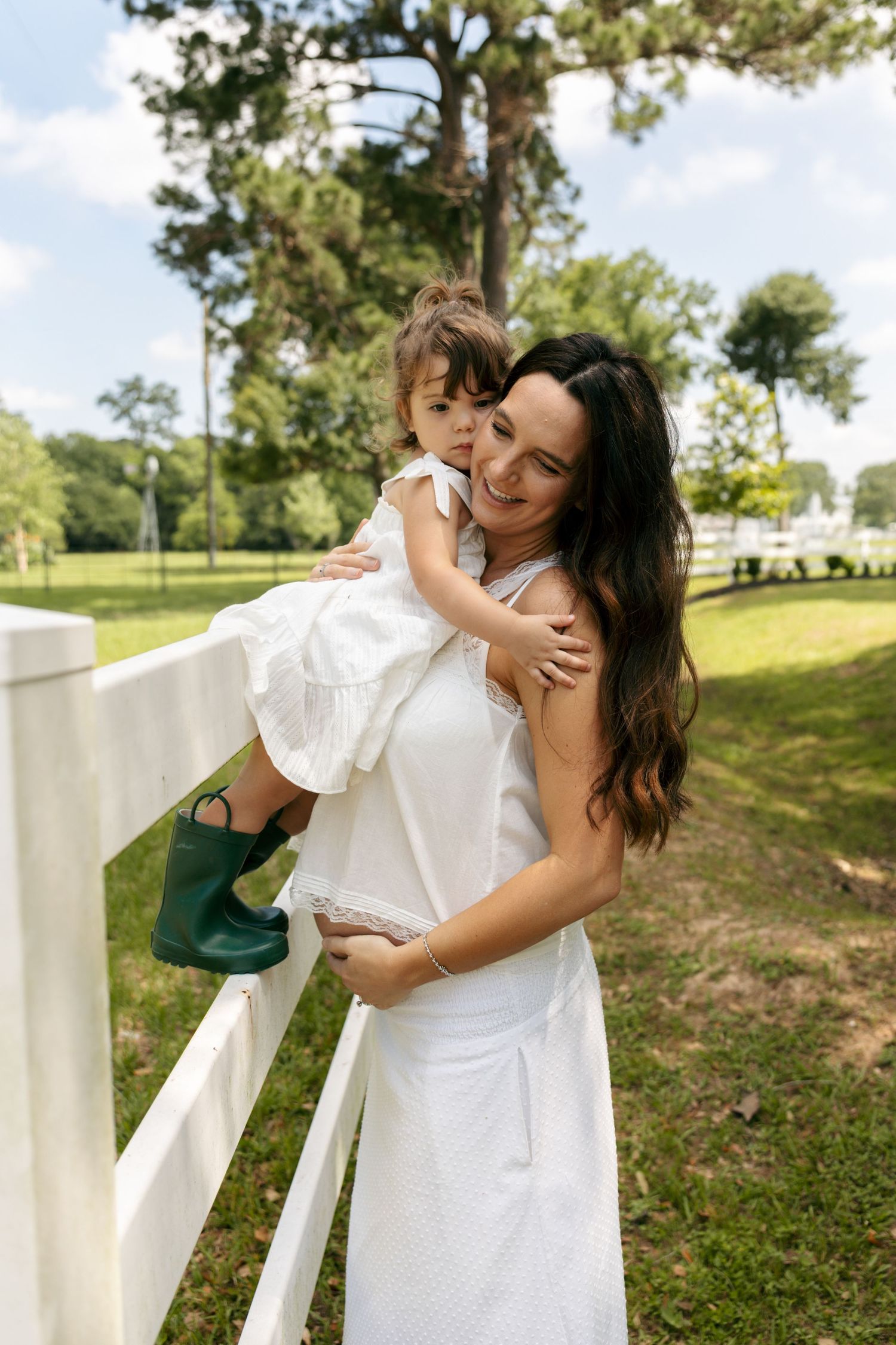 Smiling pregnant woman in white dress holds a toddler girl on a white fence outdoors.