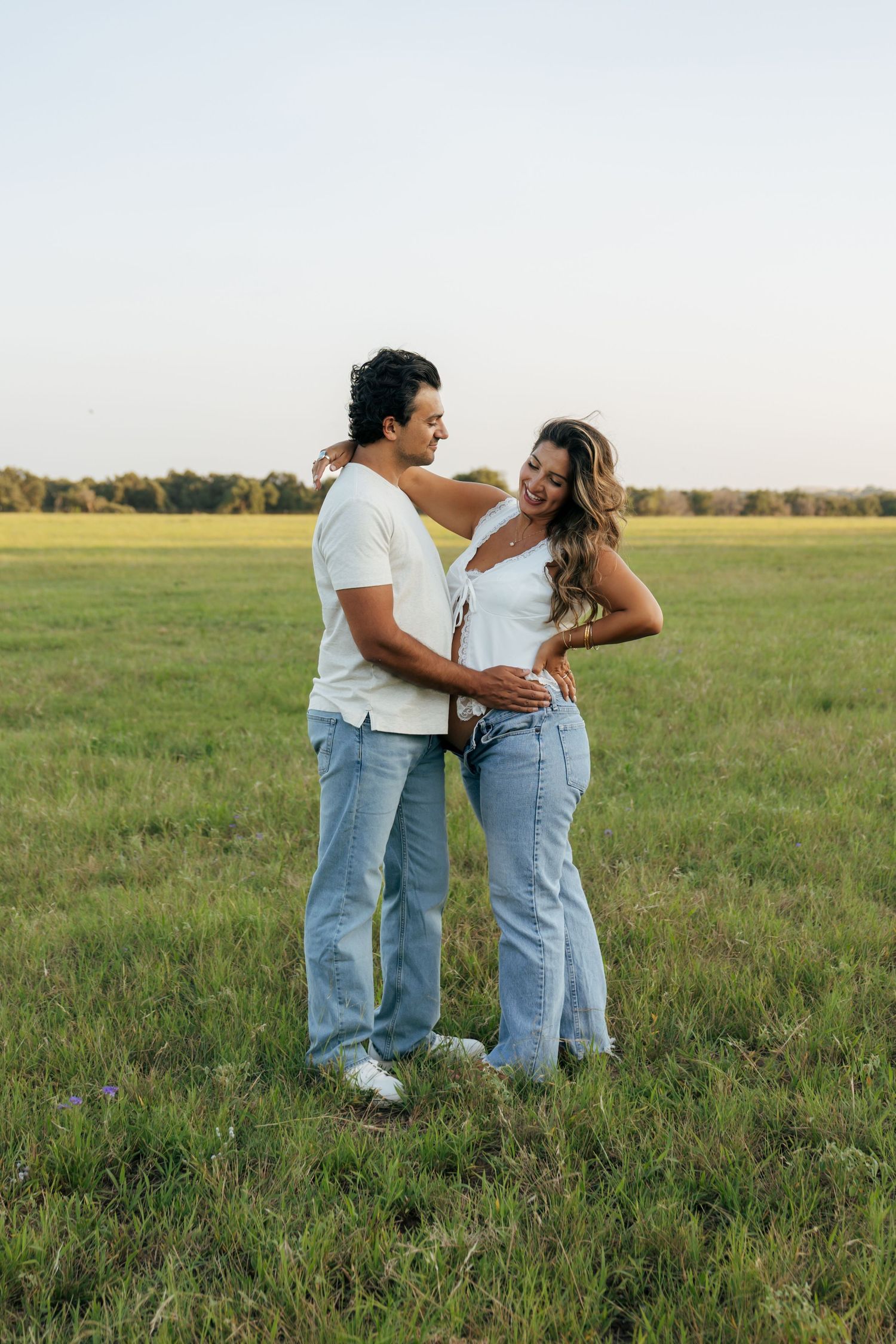 Happy couple in white tops and jeans embrace in a lush green field at sunset.