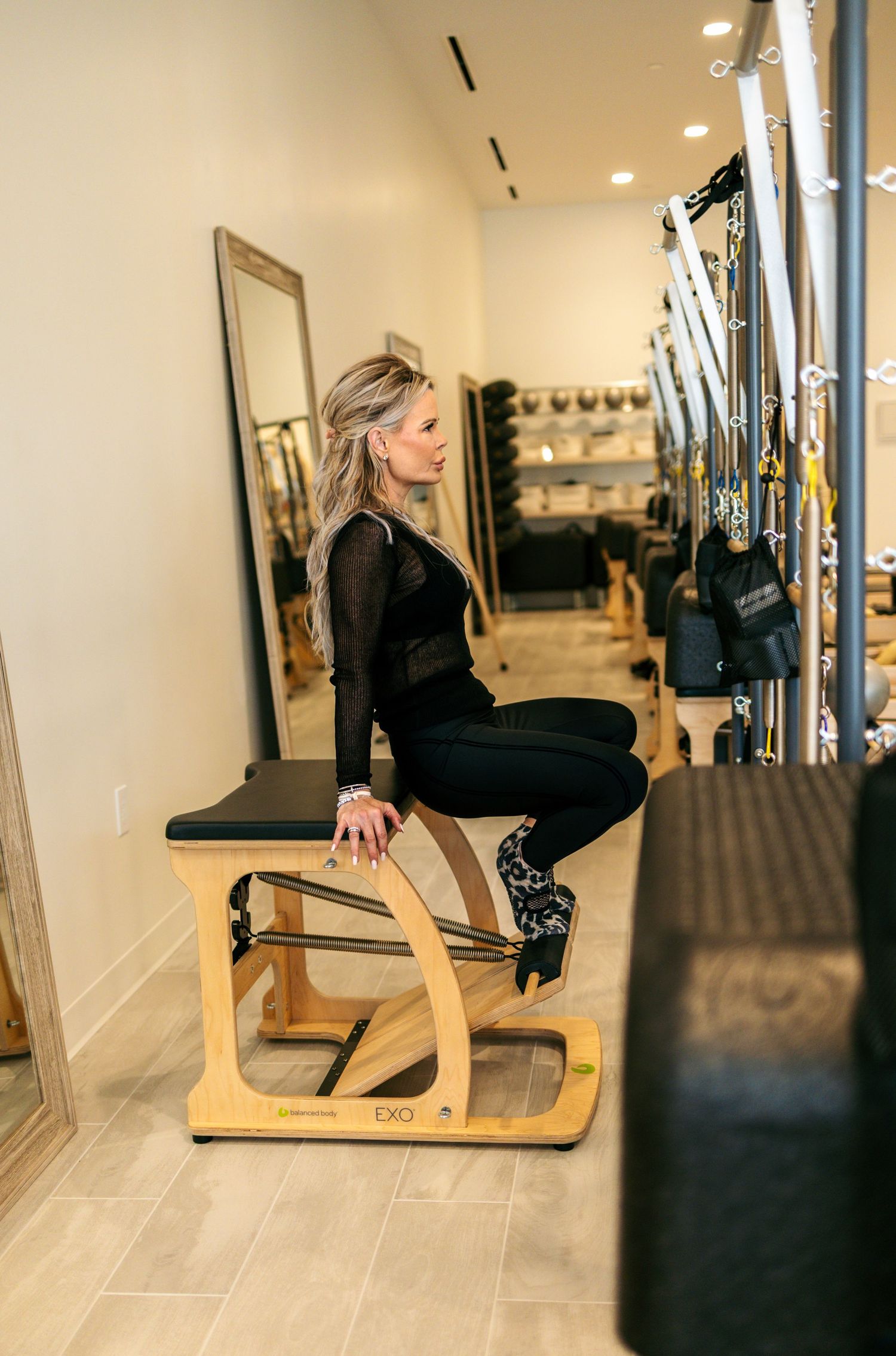 A blonde woman in black activewear sits on a wooden Pilates chair in a well-equipped studio.