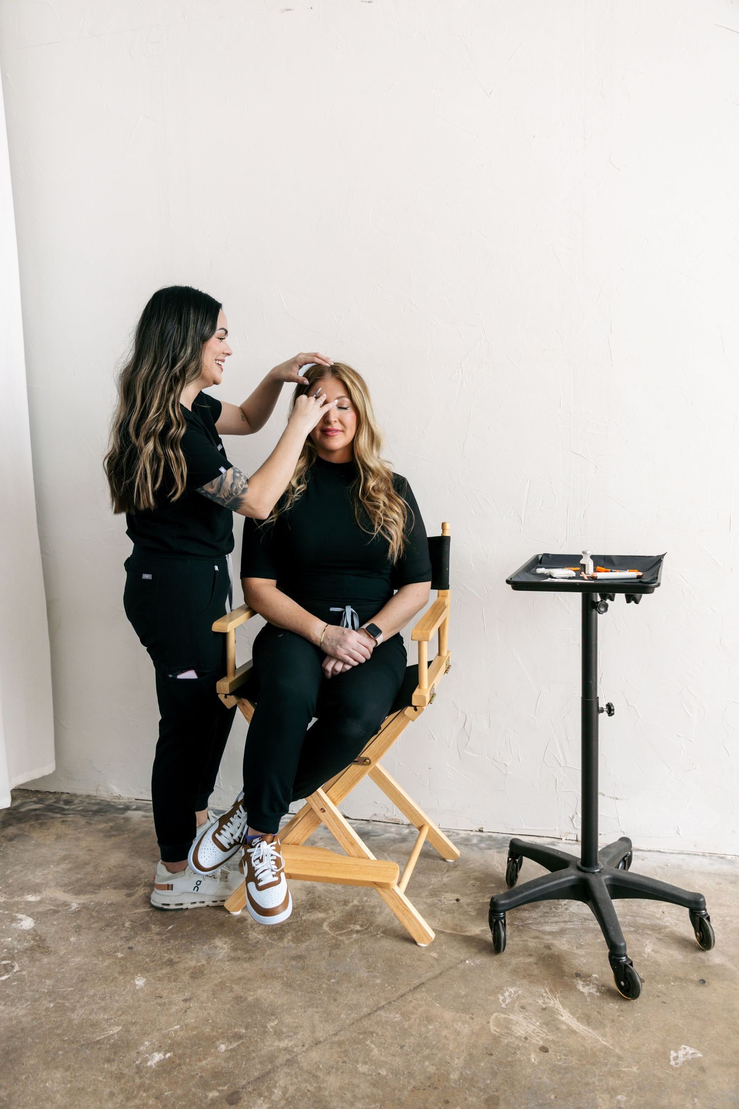 Woman in black applying makeup to another woman seated in a directors chair in a studio.
