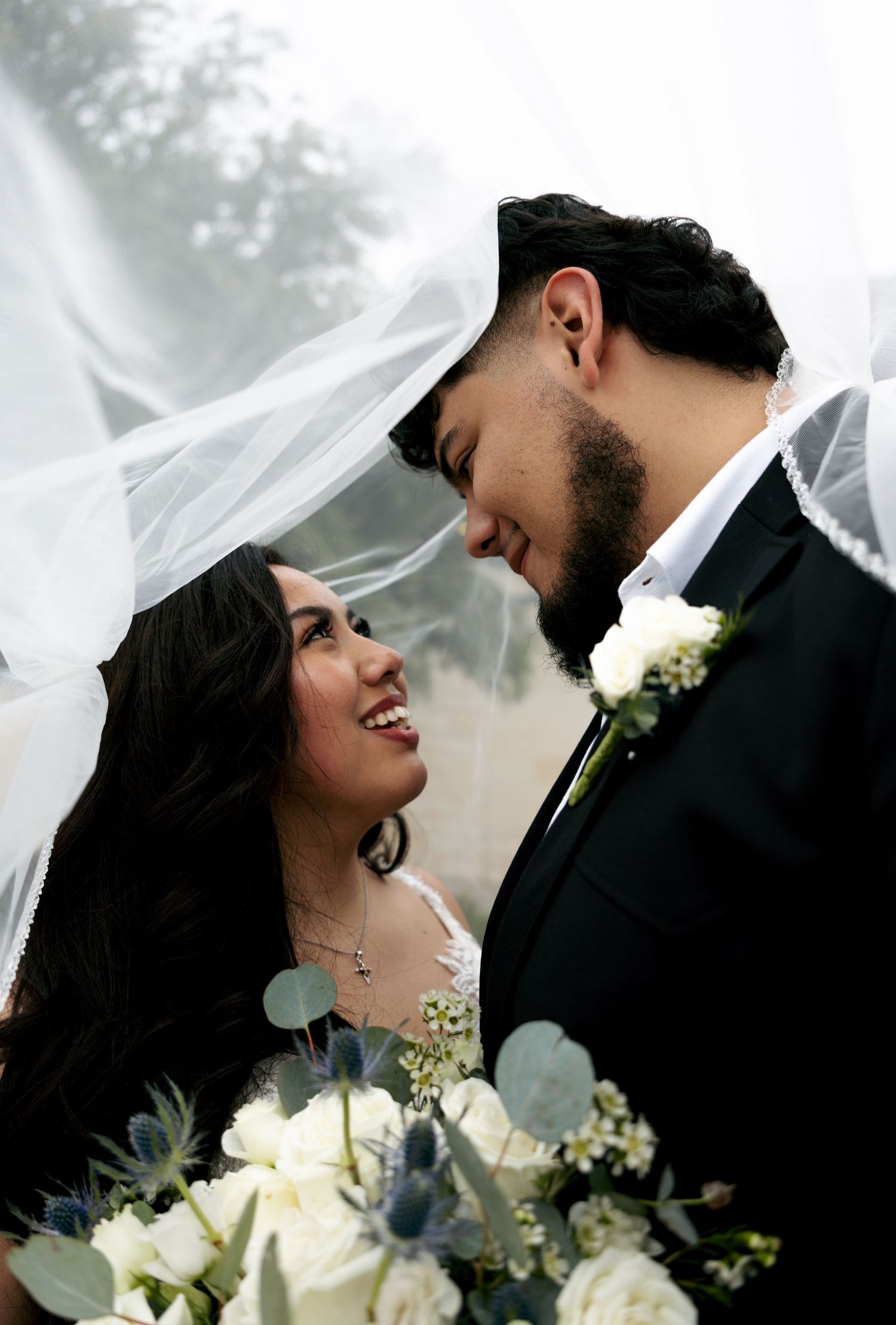 Bride and groom smiling under flowing veil with white floral bouquet.