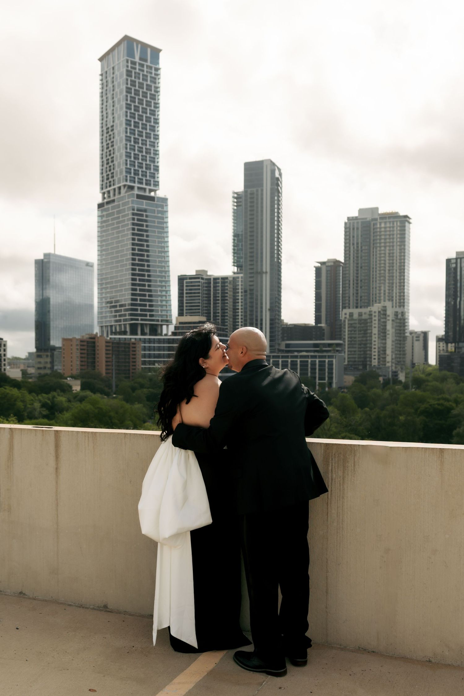 Newlyweds kissing on rooftop with city skyline backdrop on cloudy day.