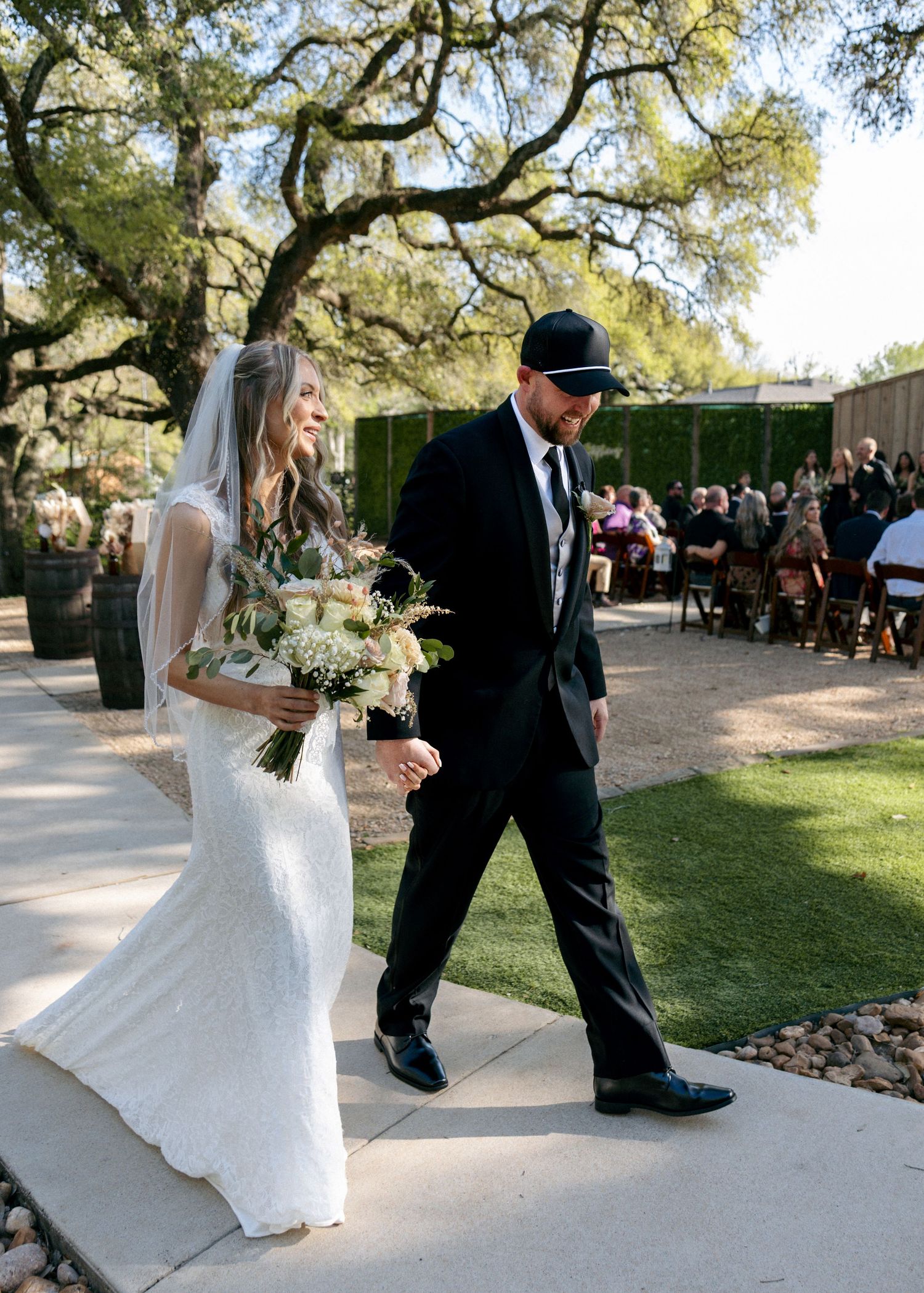 Bride in lace gown holding bouquet walks with escort in black suit and hat toward outdoor ceremony.
