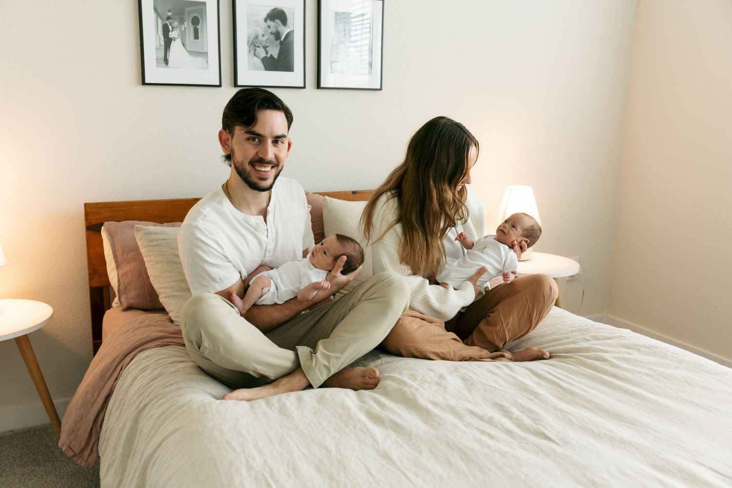 Happy couple sitting on bed holding newborn twins in a cozy bedroom.