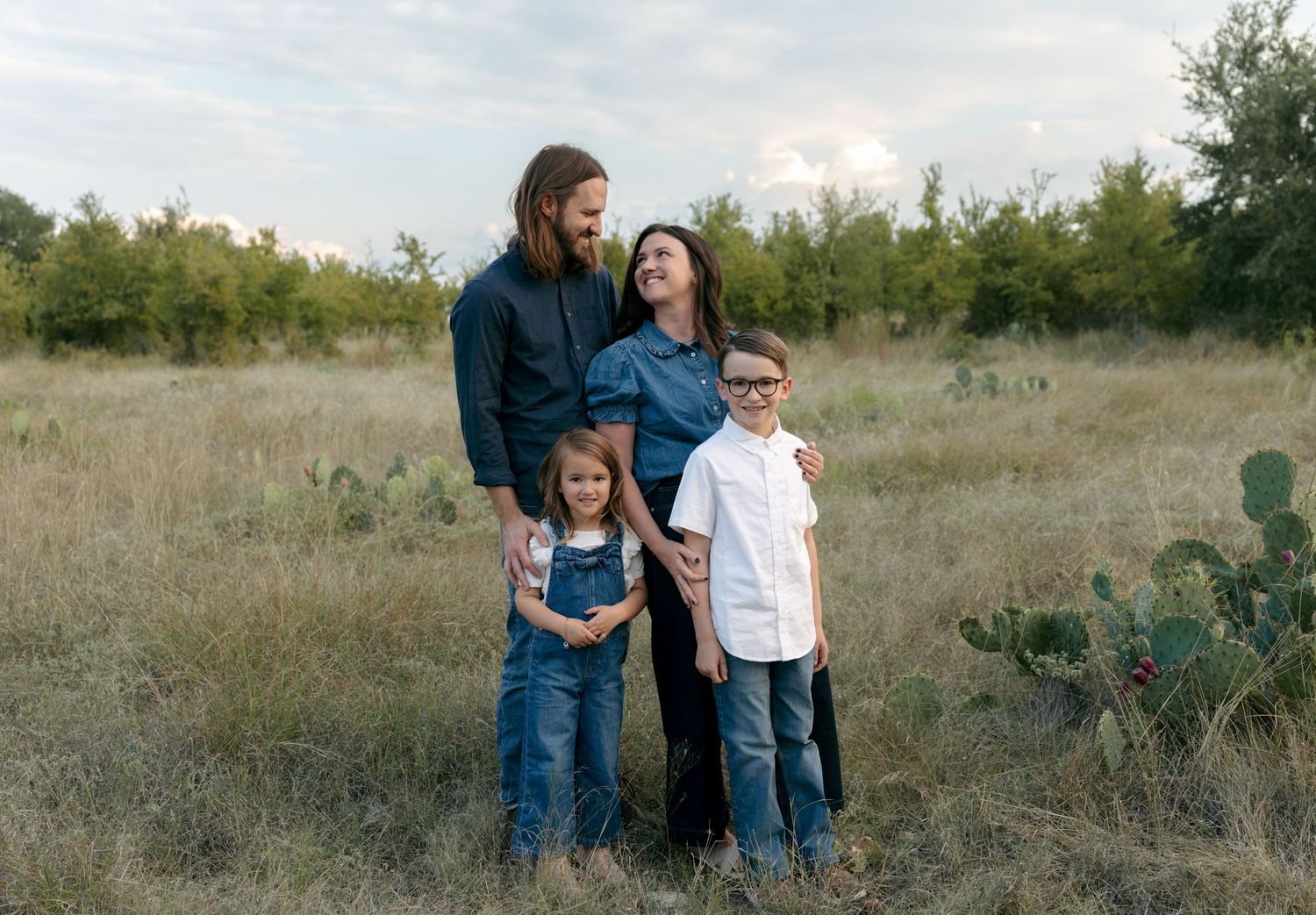 Happy family of four standing in rustic field with cactus, two children smiling with parents.