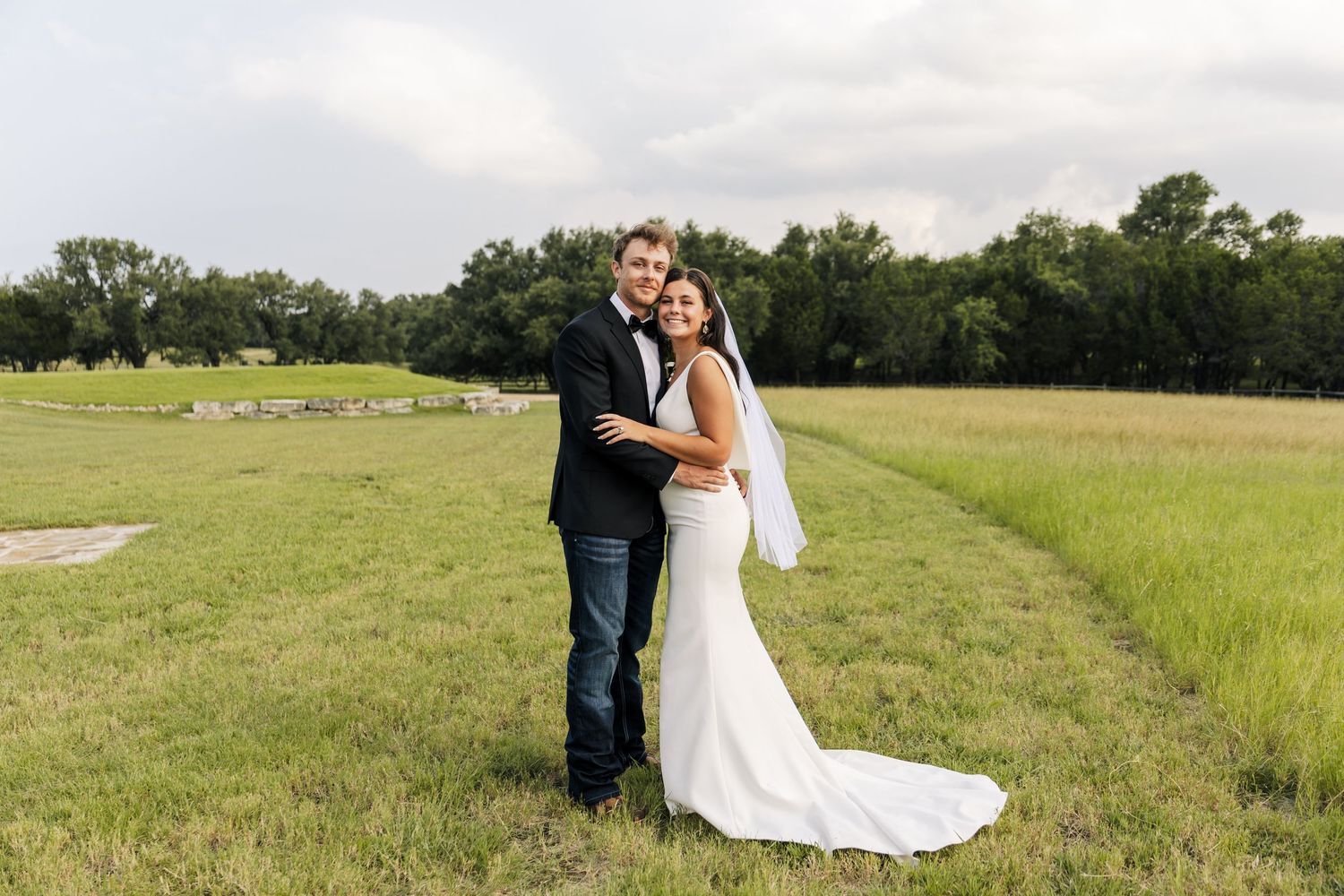 Smiling bride and groom embracing in green field, bride in fitted white gown with veil.