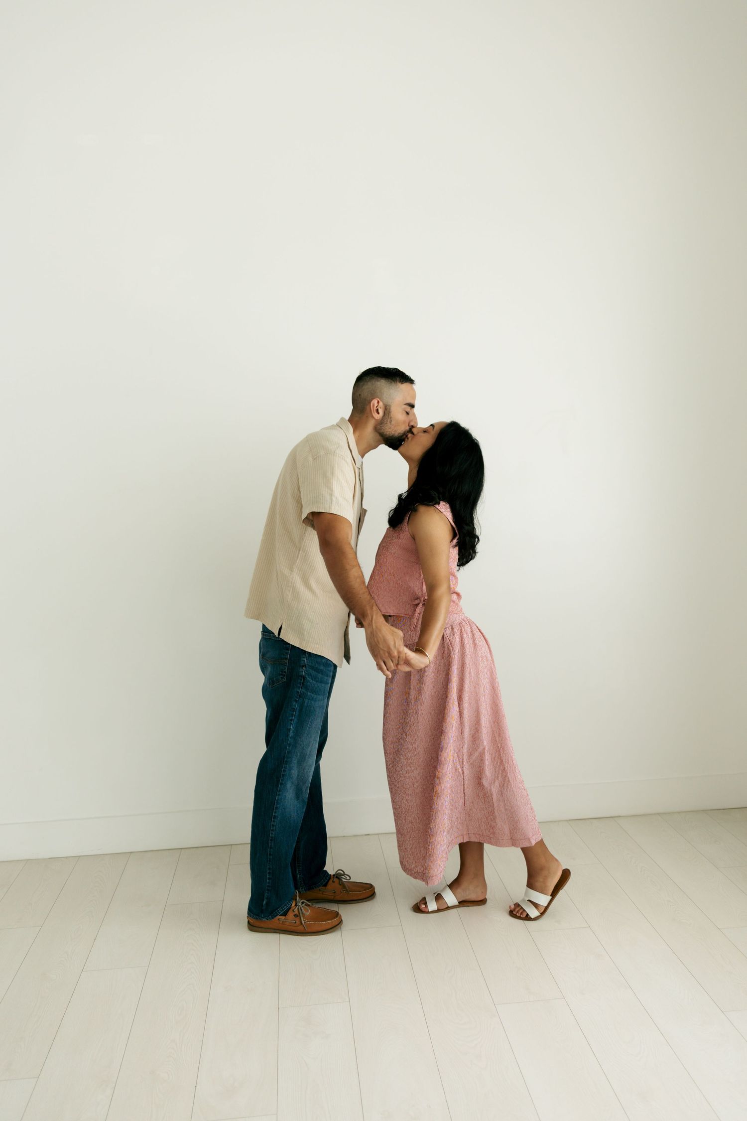 Couple sharing a kiss while holding hands in a bright white minimalist studio.