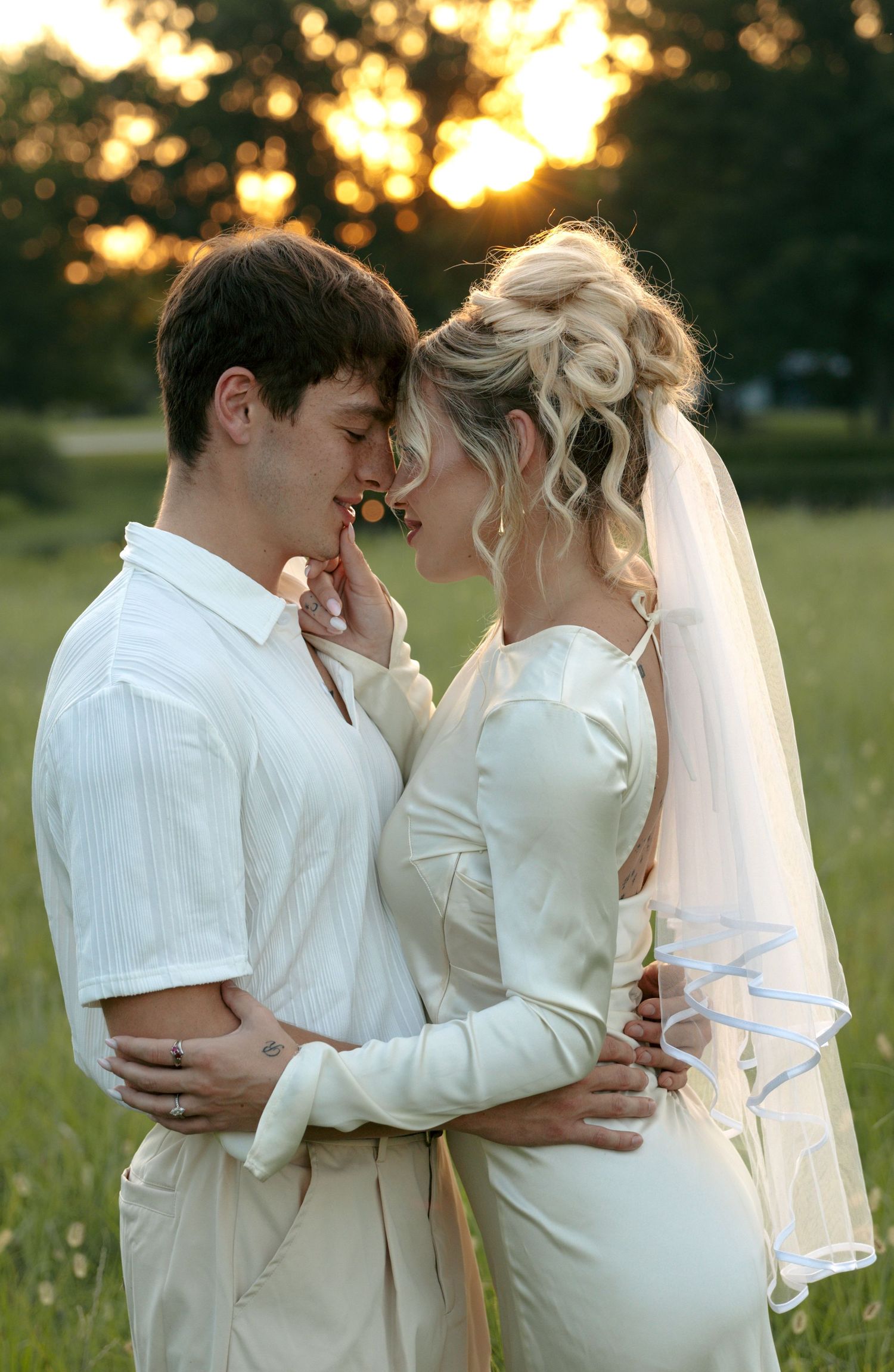 Couple in white wedding attire share a romantic forehead touch at golden hour in a field.
