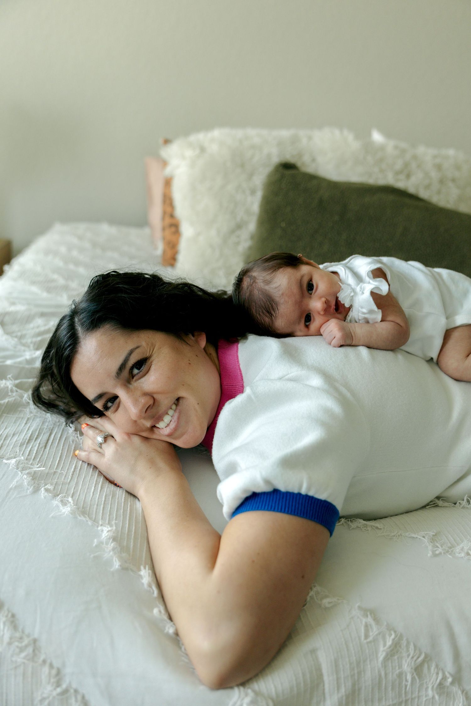 Smiling mother lying on bed with newborn baby resting on her back during an intimate home newborn photo session.