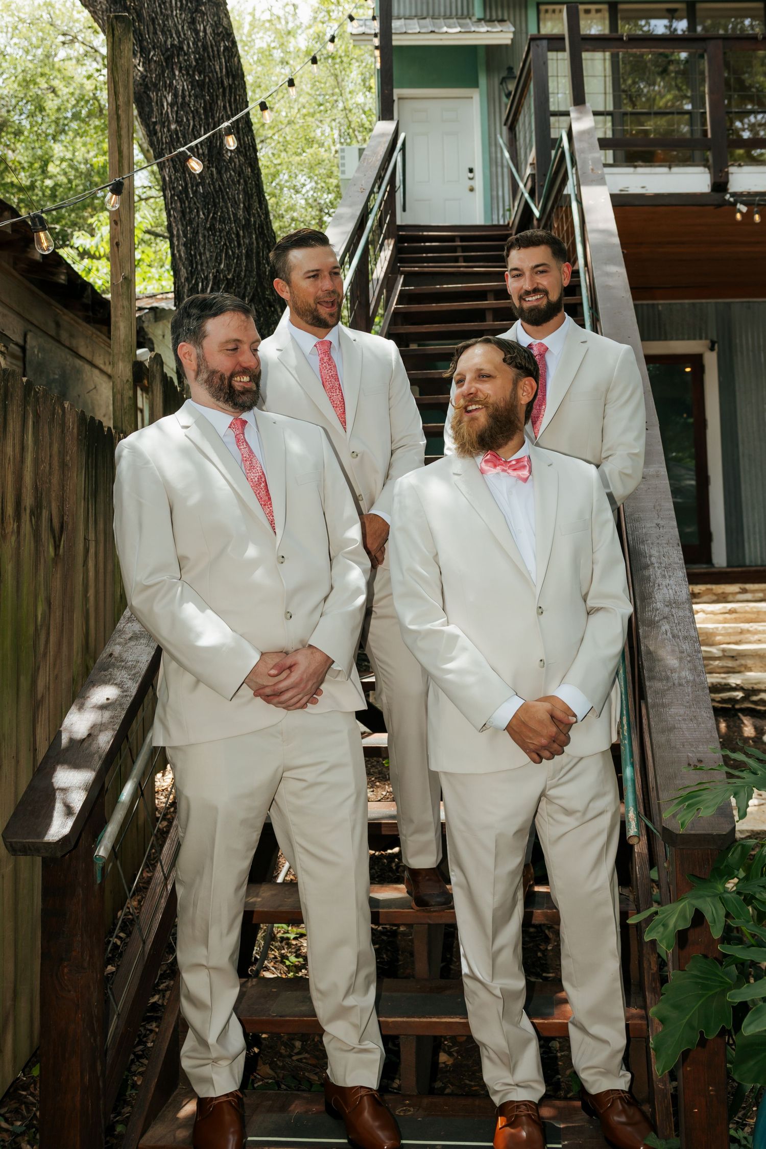 Four groomsmen in cream suits with pink ties posing on wooden stairs outdoors.