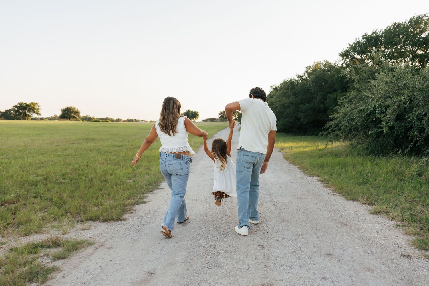 Family of three walking down a country dirt path surrounded by green fields at dusk.